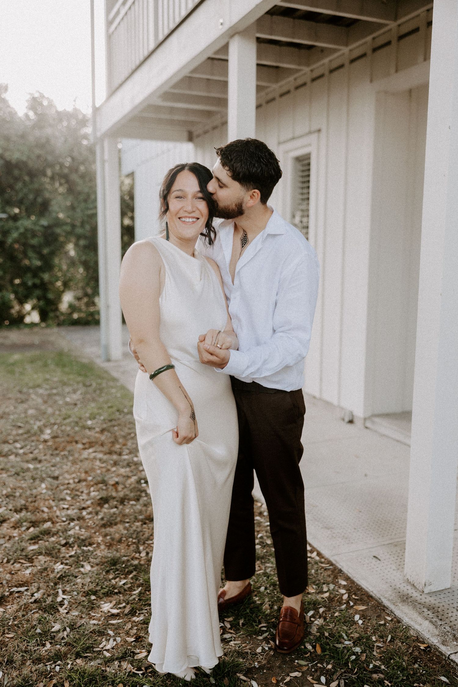 A romantic couple poses together on a white porch, wearing elegant white and neutral attire.