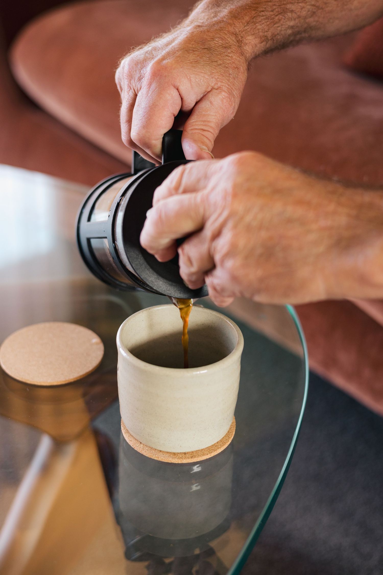 Hands pouring fresh coffee from an aeropress into a ceramic cup on glass table.