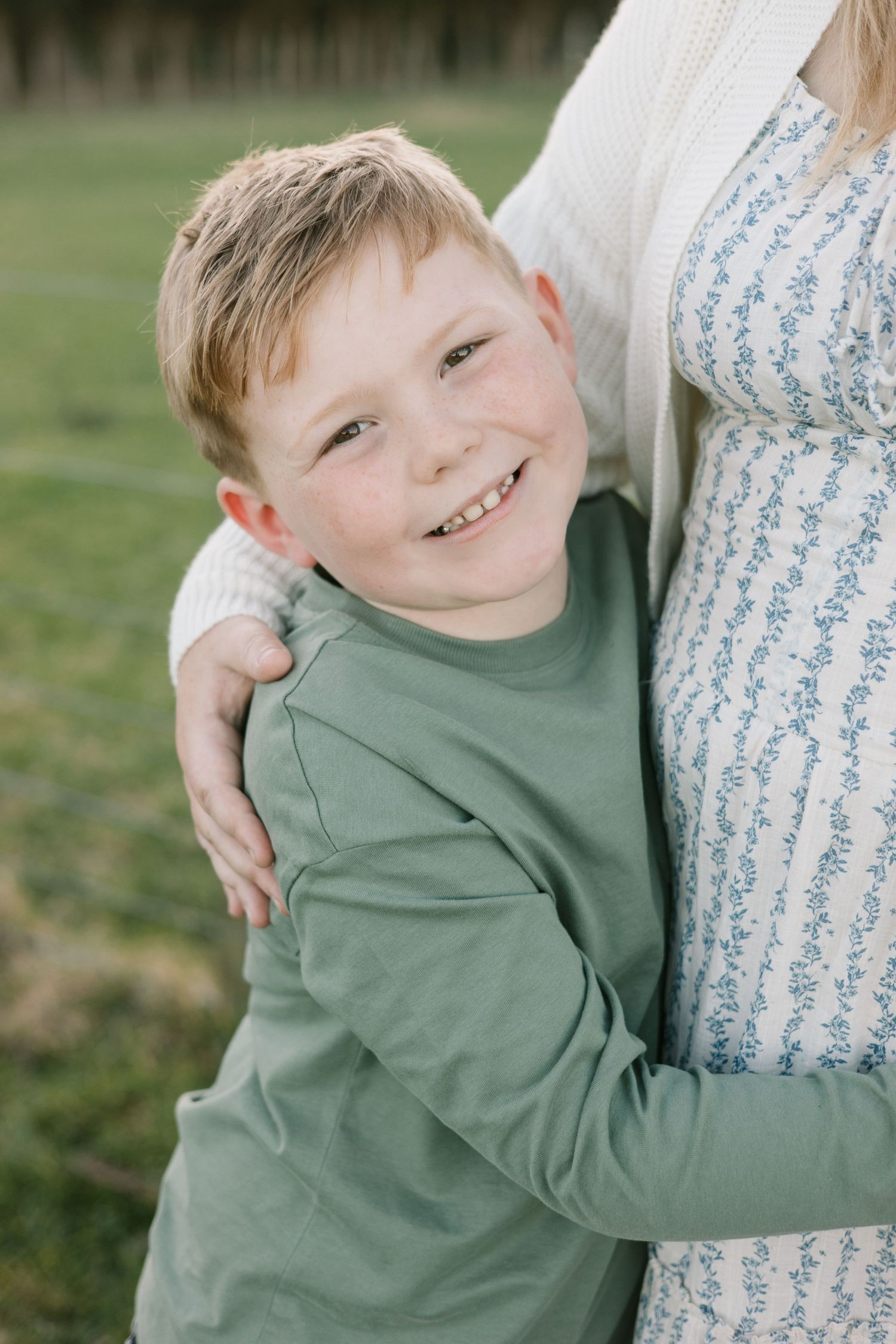 A joyful embrace captured outdoors with a green sweater and floral dress visible in the frame.