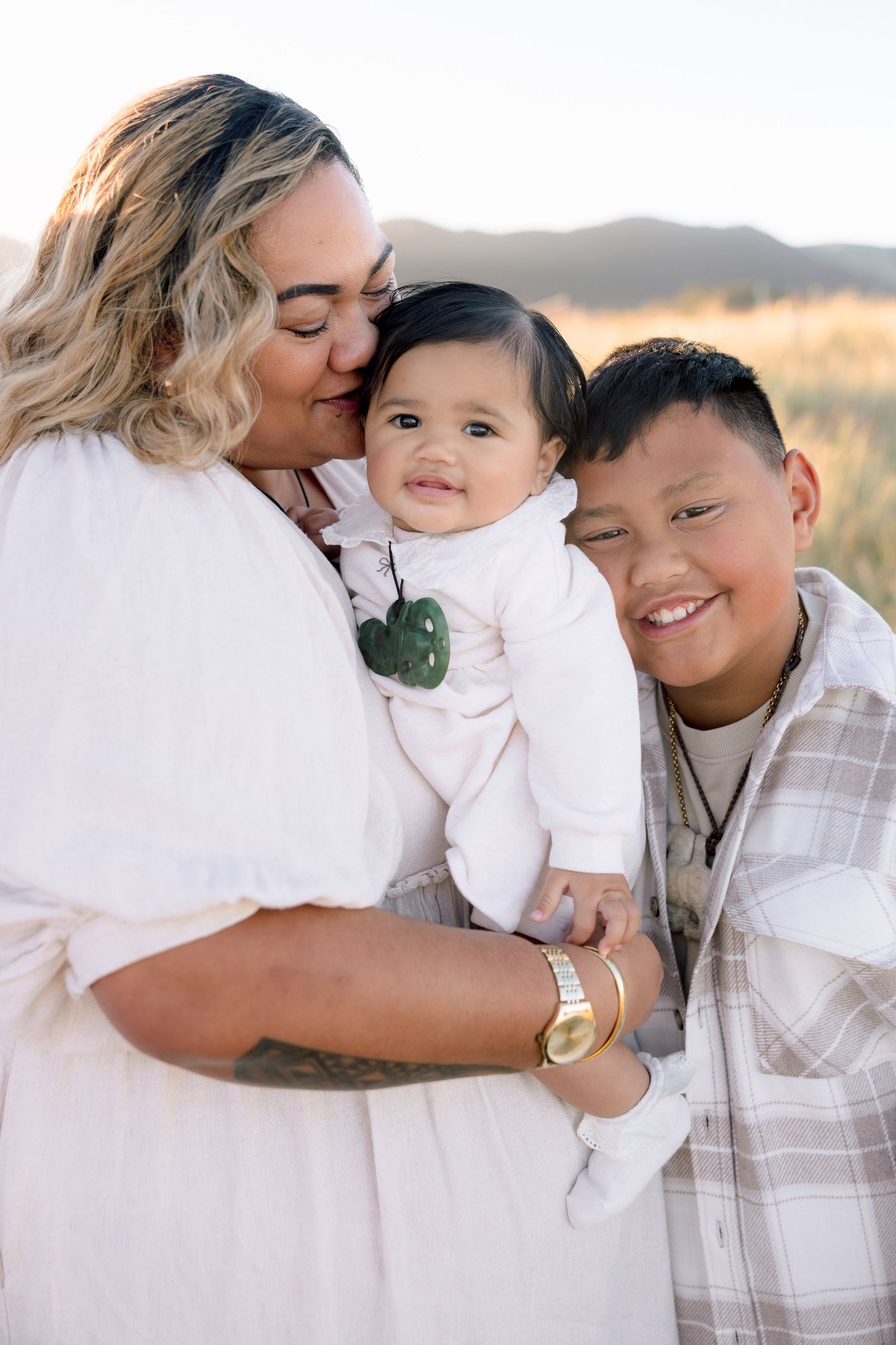 A mother kisses her smiling baby while her young son smiles beside them in a sunny outdoor field.