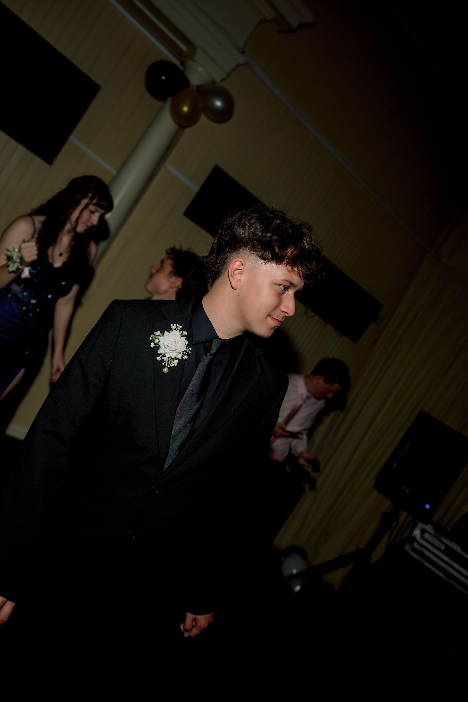Young man in black suit with white boutonniere at a formal prom event with balloons in background.