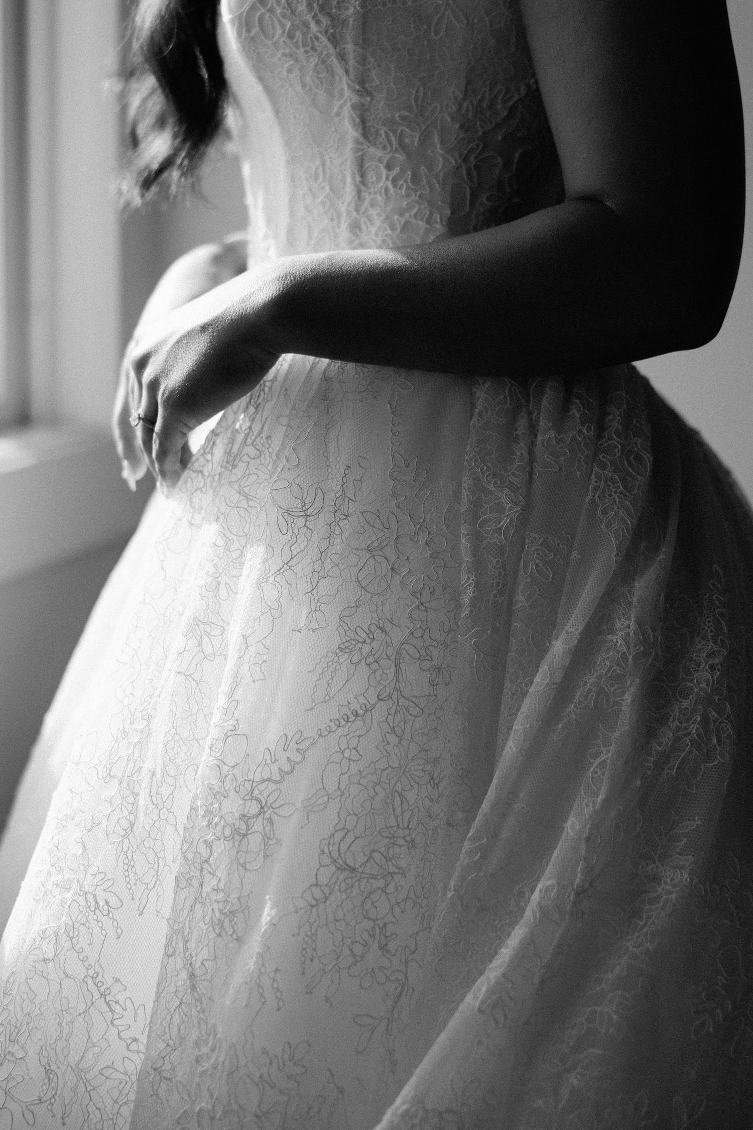 Black and white photo of a bride in an elegant lace wedding gown standing by a window, arms gently crossed.