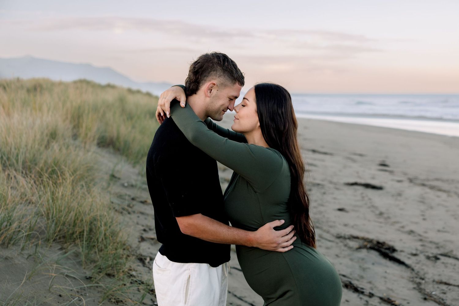 Couple embracing on a sandy beach at sunset, woman in green dress and man in black shirt sharing a romantic moment.