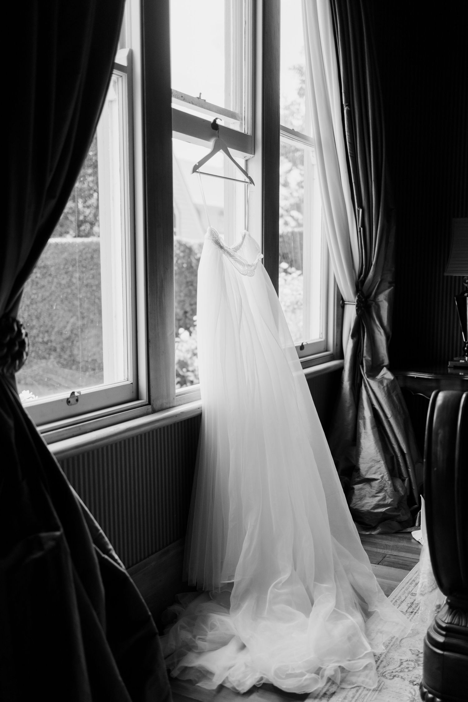 A white wedding dress hangs elegantly by a window with dark curtains in black and white photography.