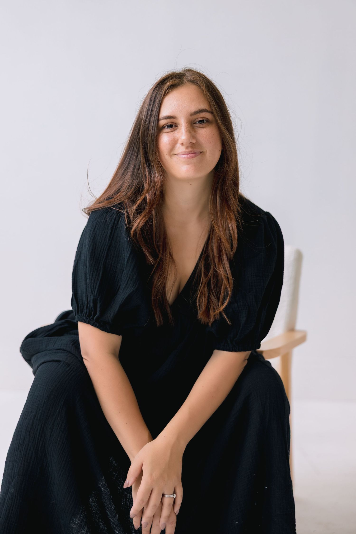 A person in a black dress sits casually against a white background with long brown hair and a warm expression.
