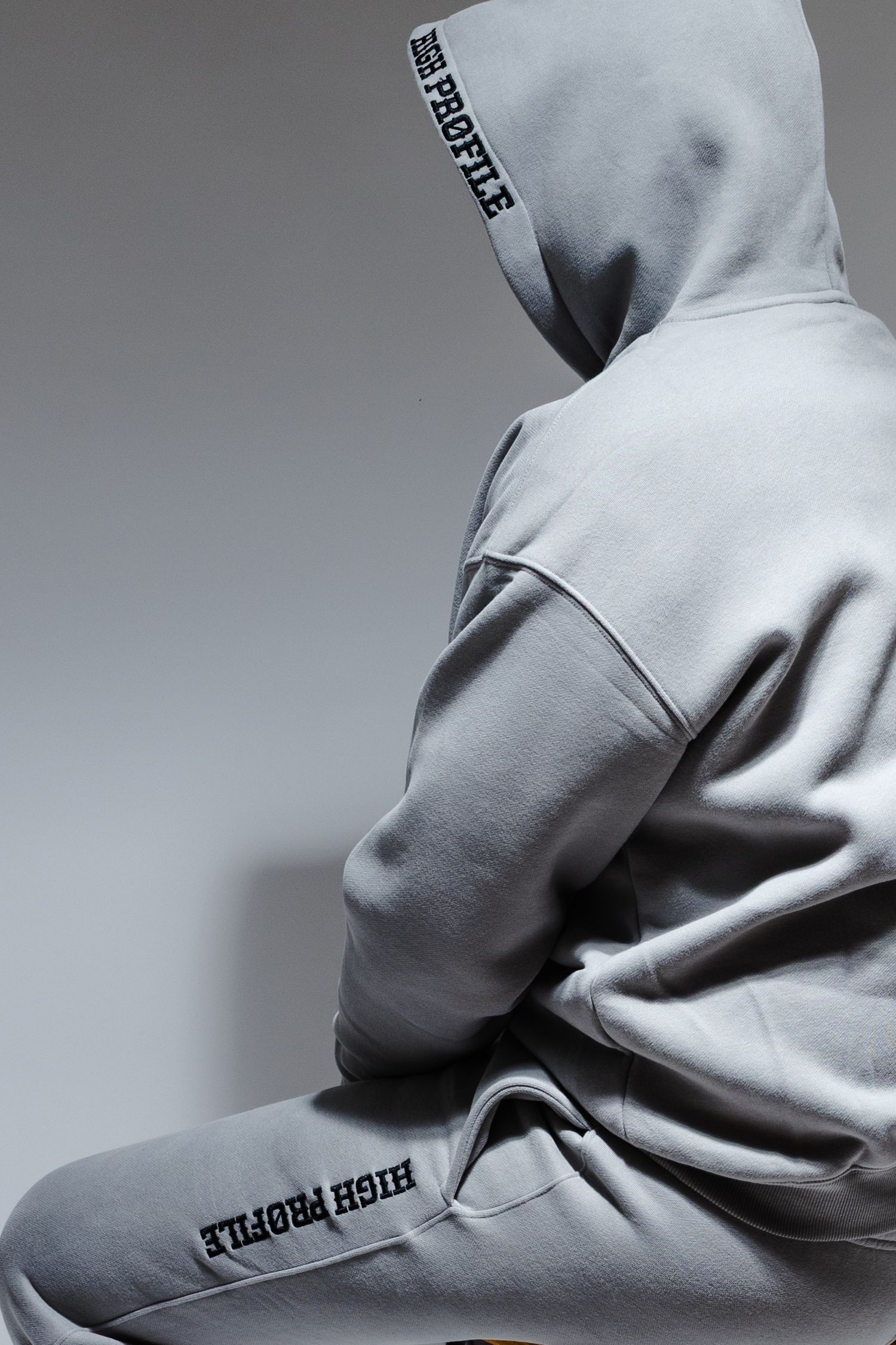 Person wearing a light gray hooded sweatsuit with High Profile text detail shown from behind against gray background.