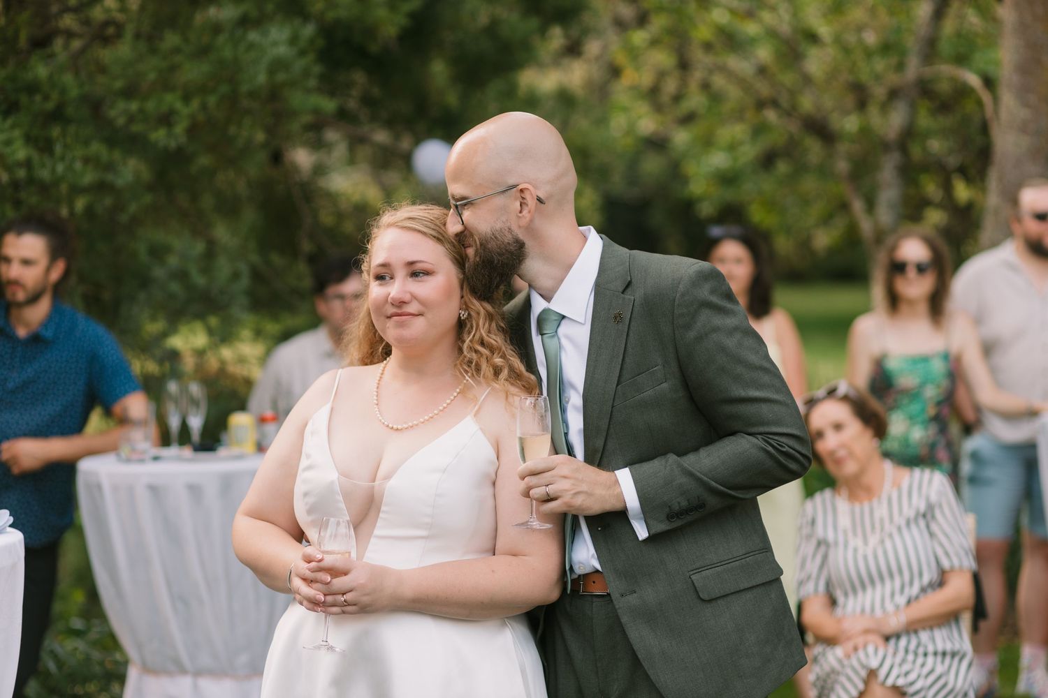 Couple shares an intimate moment during outdoor wedding reception with guests in background.
