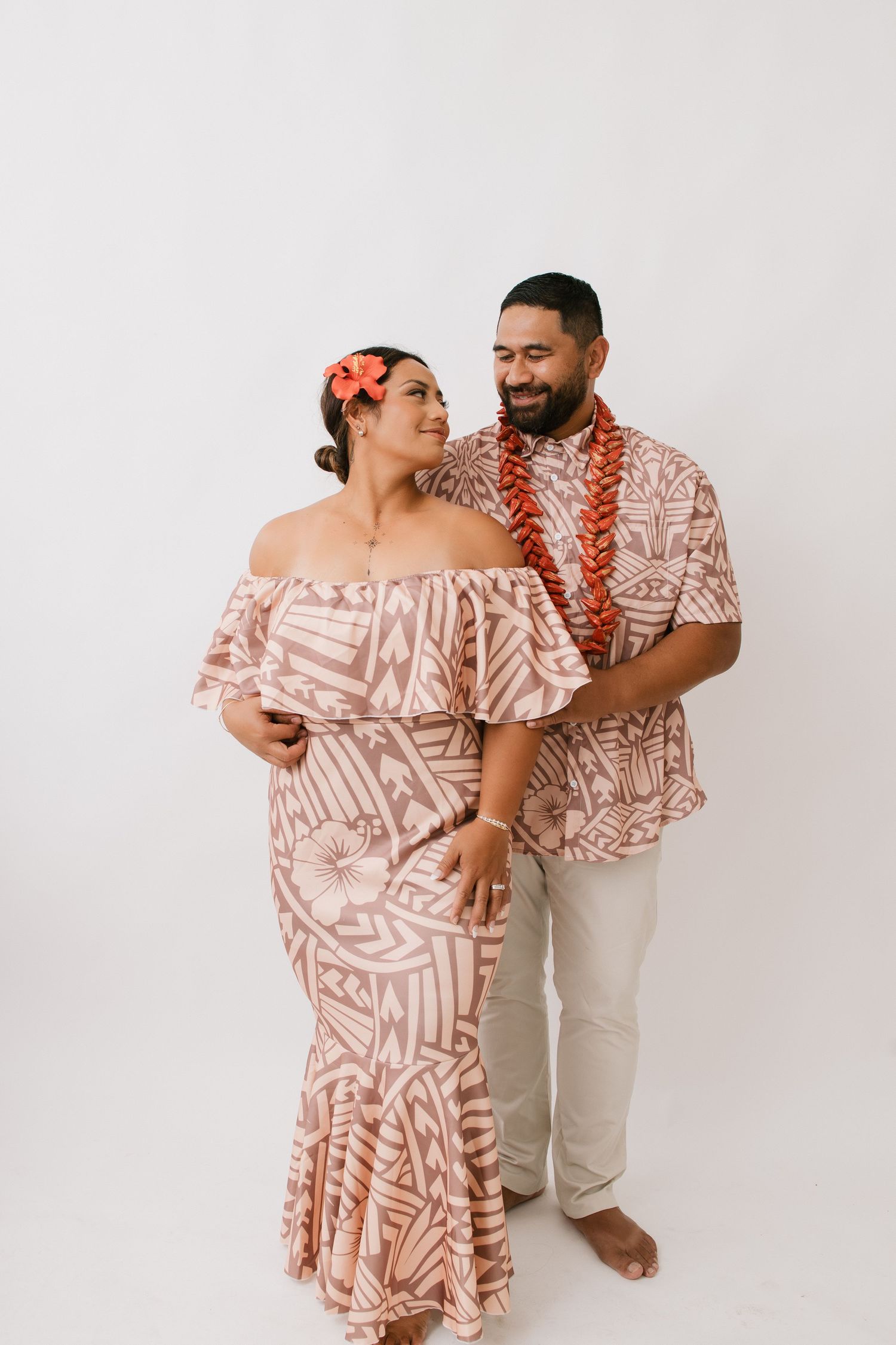 Couple wearing matching Hawaiian-style outfits with pink floral patterns embrace and laugh against white background.