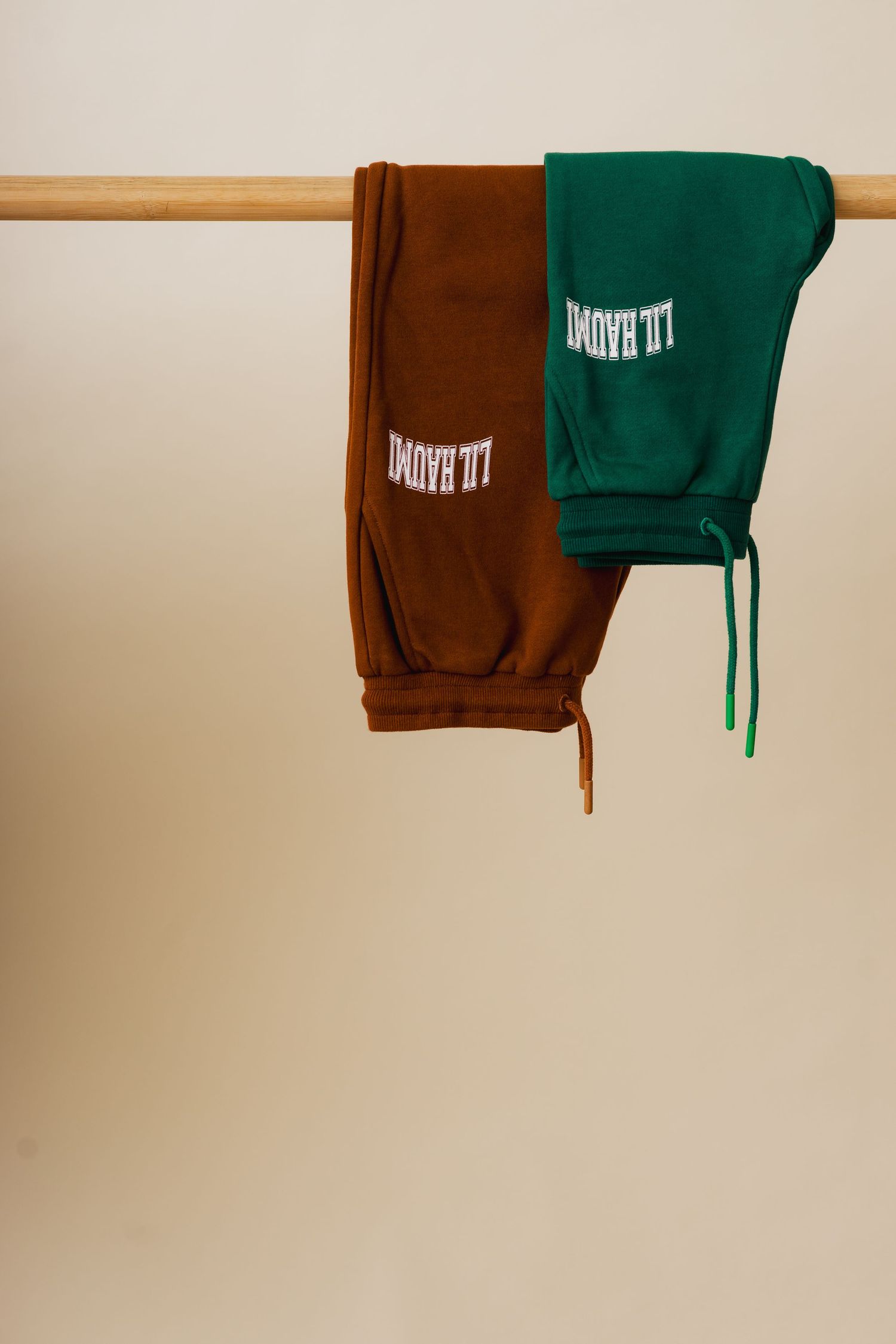 Two sweatshirts, one brown and one green, hanging side by side on a wooden clothing rod against a beige wall.