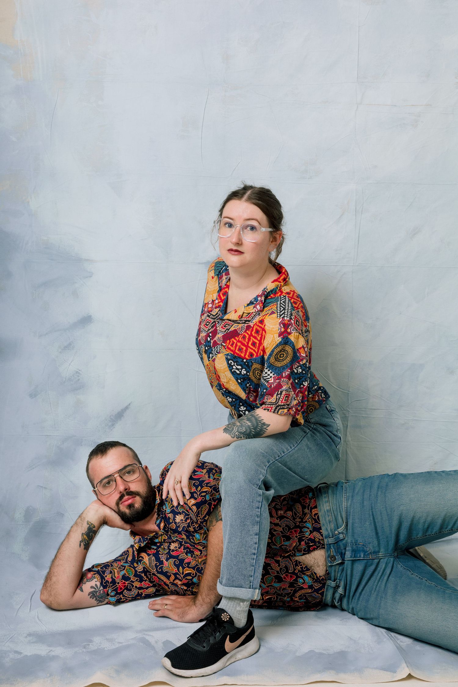A couple in colorful patterned shirts and jeans pose casually against a light gray backdrop during a studio photoshoot.