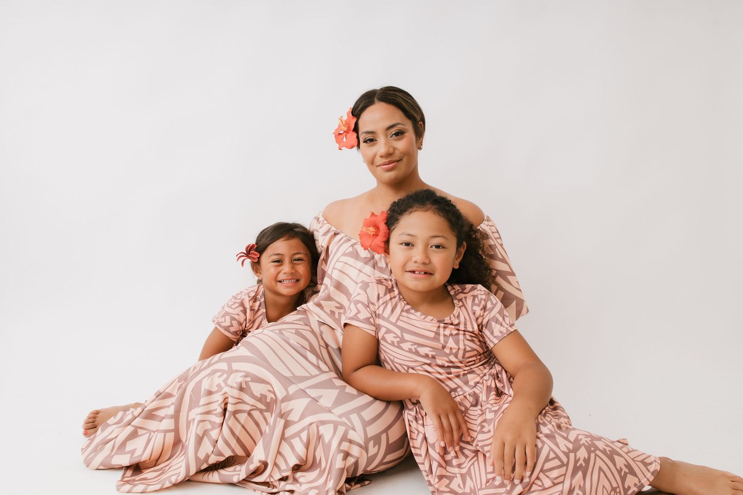 Three people wearing matching pink and white patterned dresses pose together on a light backdrop for a family portrait.