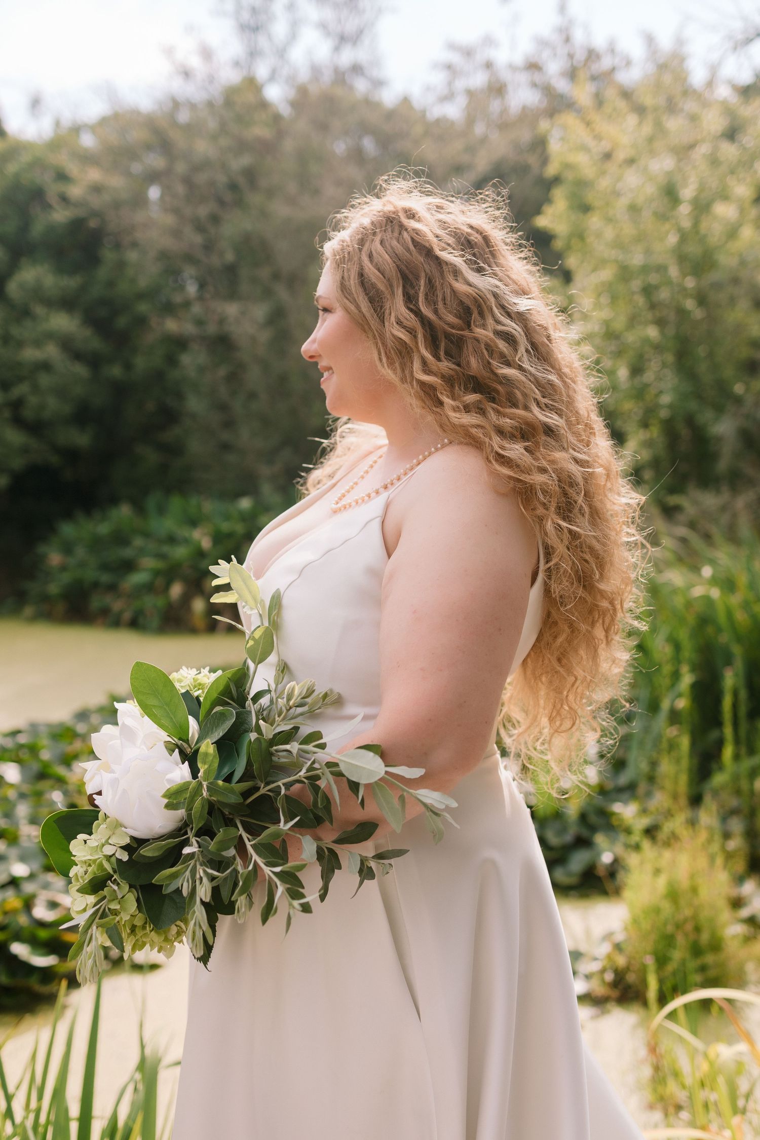A bride in a sleeveless white gown holds a lush bouquet of white flowers and greenery in a garden setting.