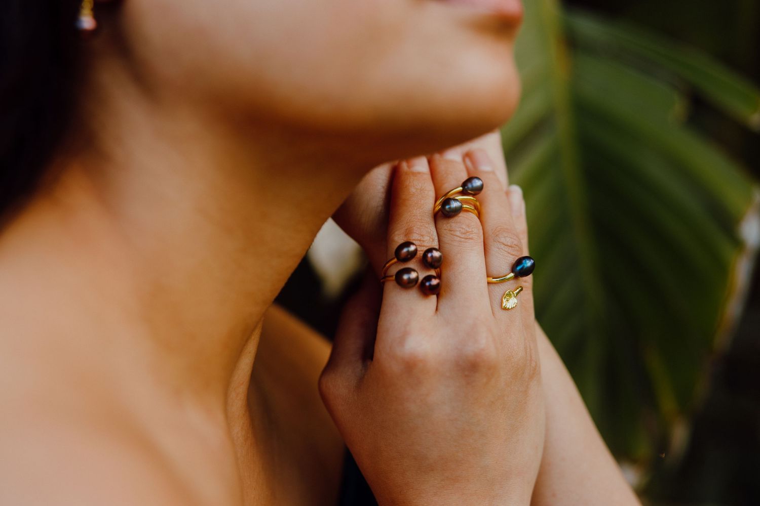 Close up of hands wearing multiple delicate rings against green palm leaves in soft natural lighting.