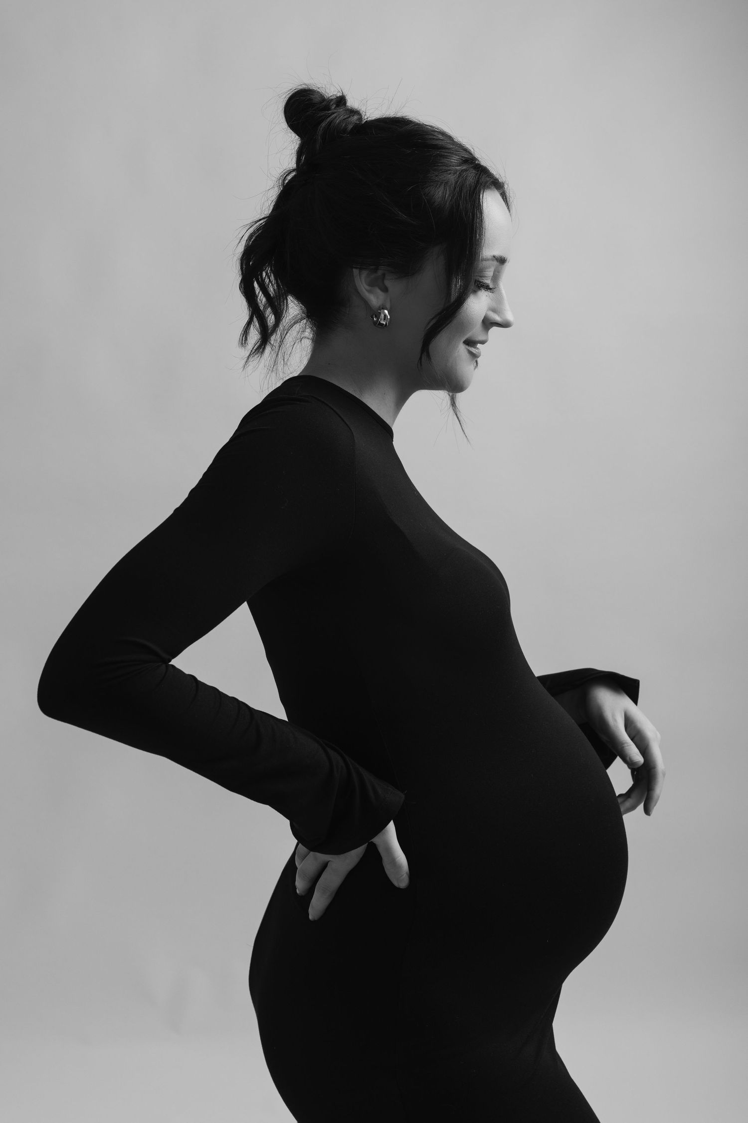 Black and white side profile of a smiling pregnant woman in a black fitted dress, hand on her baby bump.
