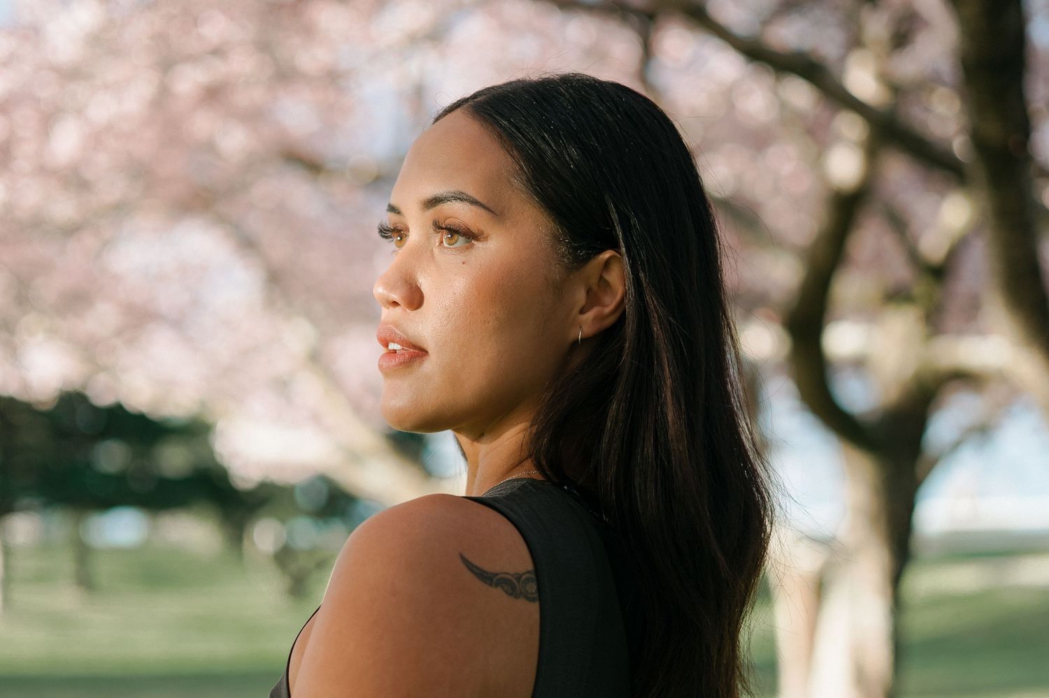A woman with long dark hair gazes into the distance with cherry blossoms blooming in the background.