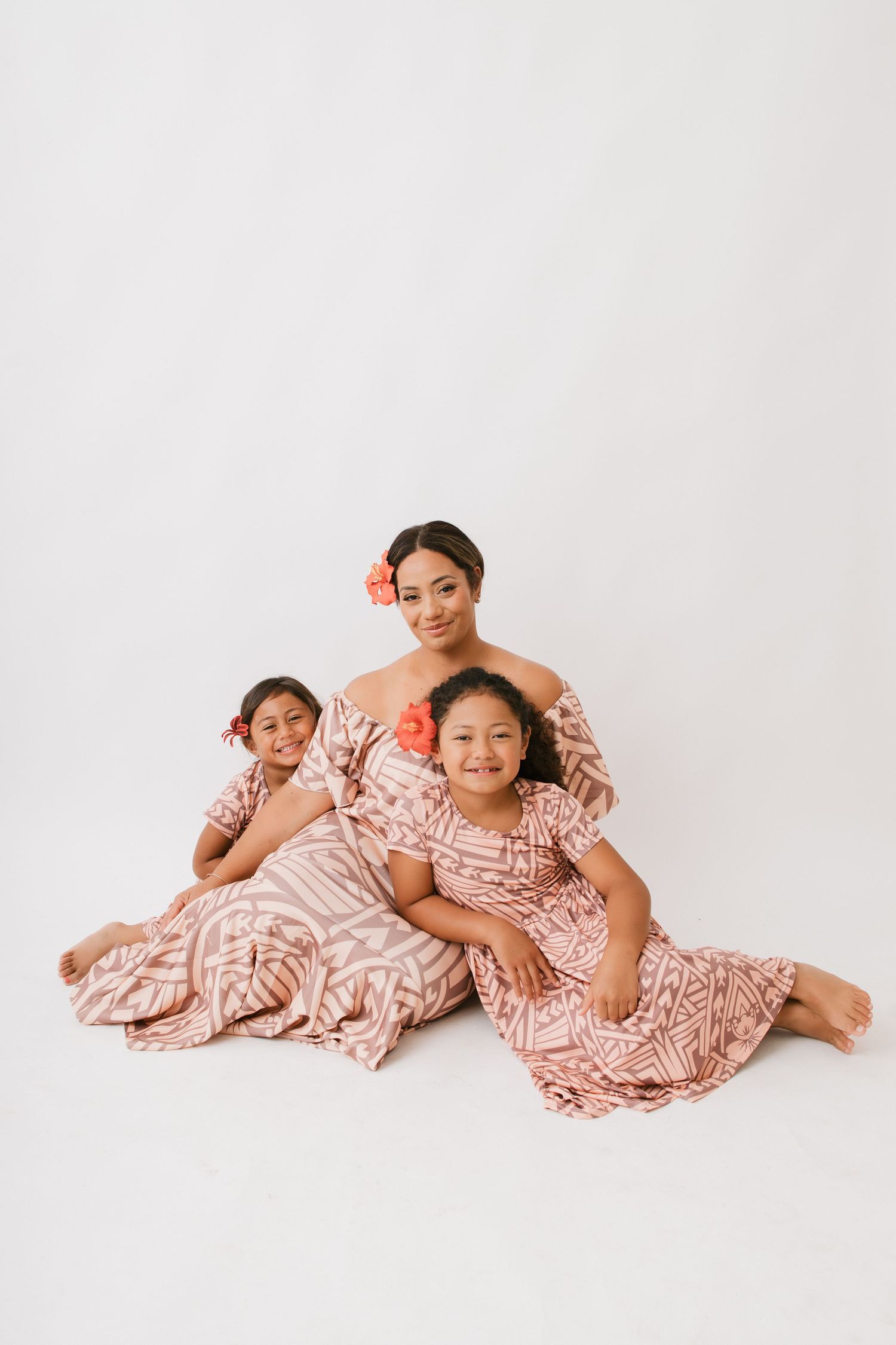 Three people wearing matching pink and white patterned dresses lay casually on a white studio backdrop for a portrait.