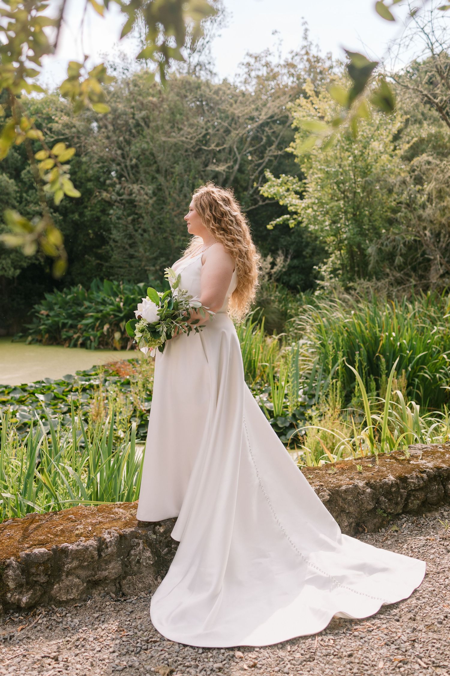 Bride in flowing white dress with train poses in lush garden setting holding a bouquet.