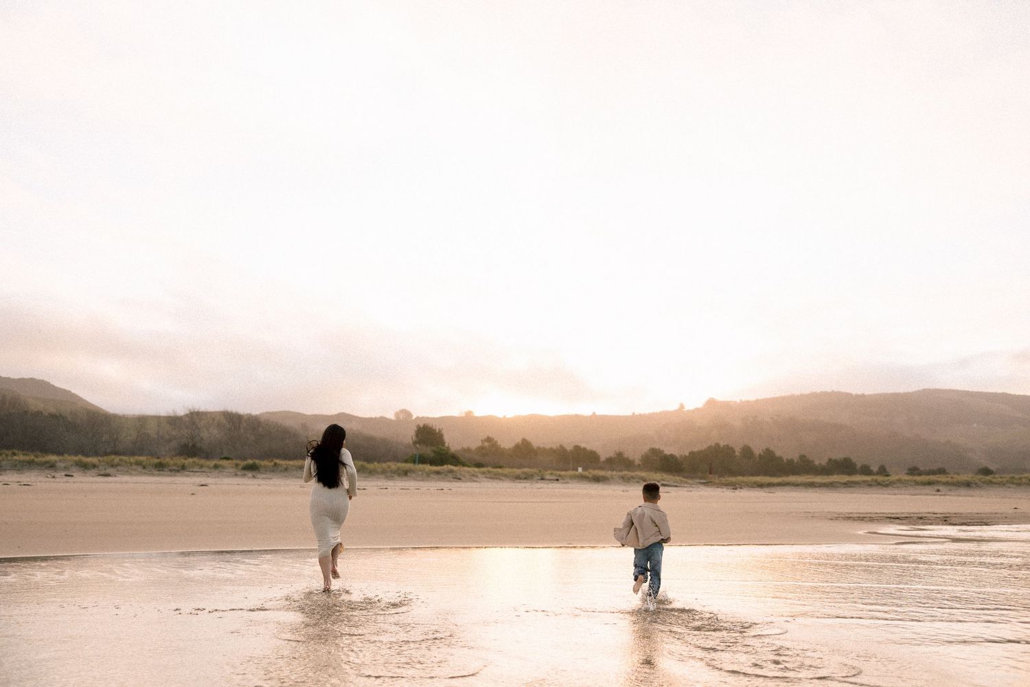 Woman and child running on a sunlit beach with mountains in the background at golden hour.