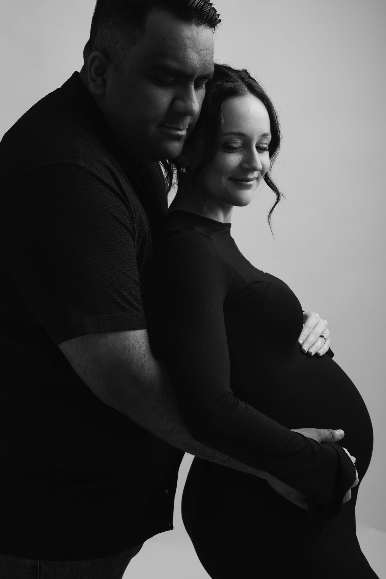 Black and white maternity photo of a couple, man embracing pregnant woman from behind, both looking down tenderly.