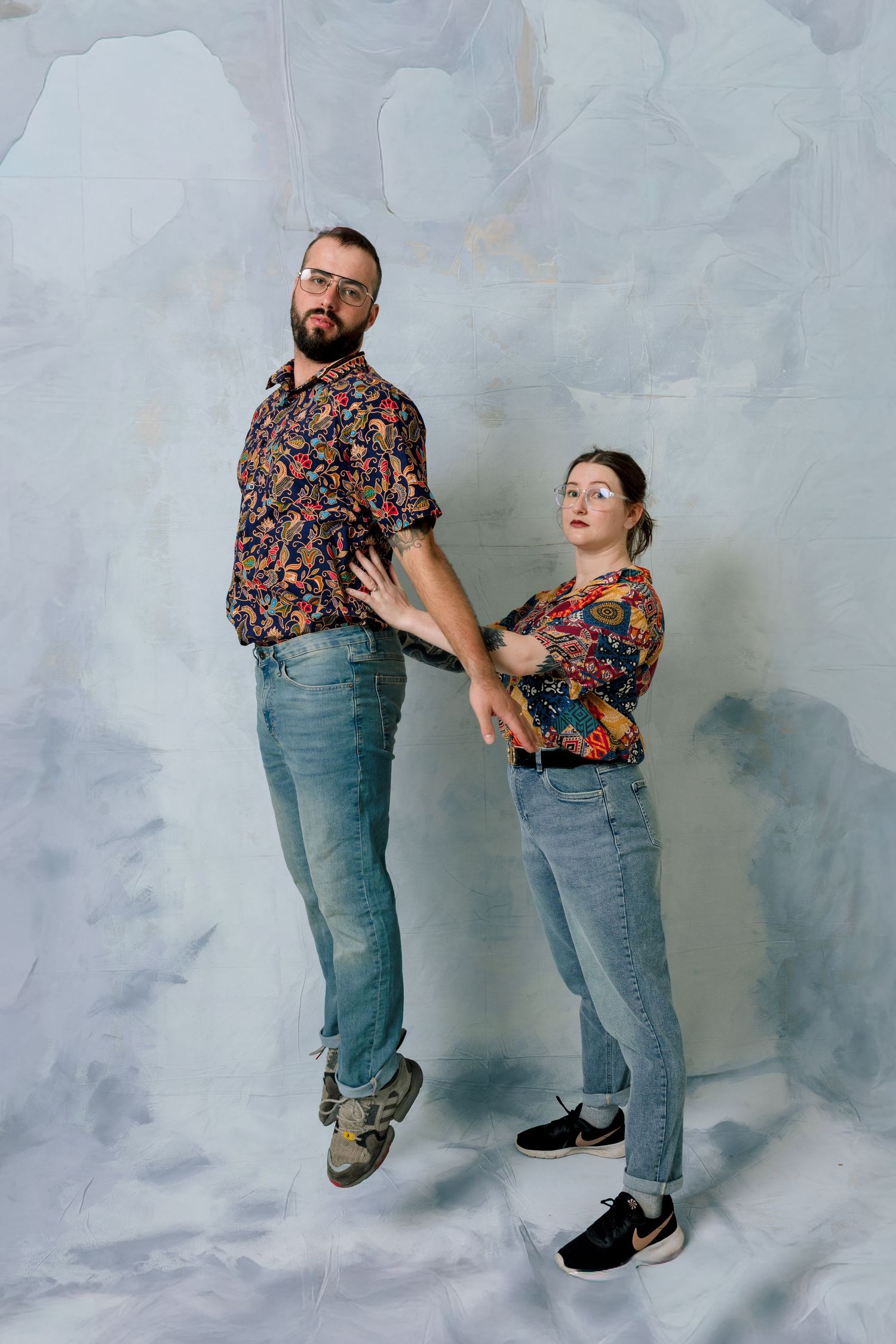 Two people in matching floral shirts and blue jeans pose playfully against a gray background.