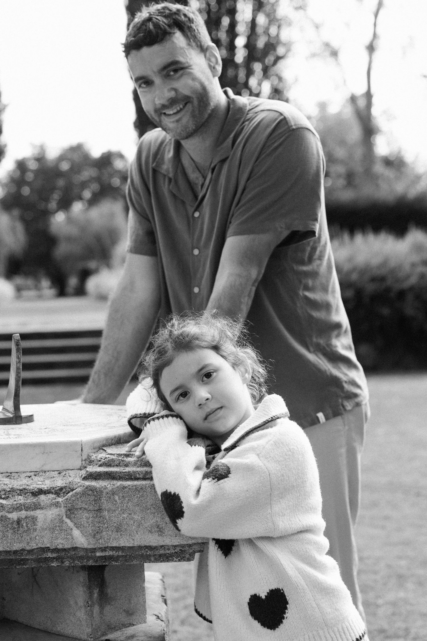 Black and white photo of a smiling man and young girl in heart sweater posing by a stone structure in a park.