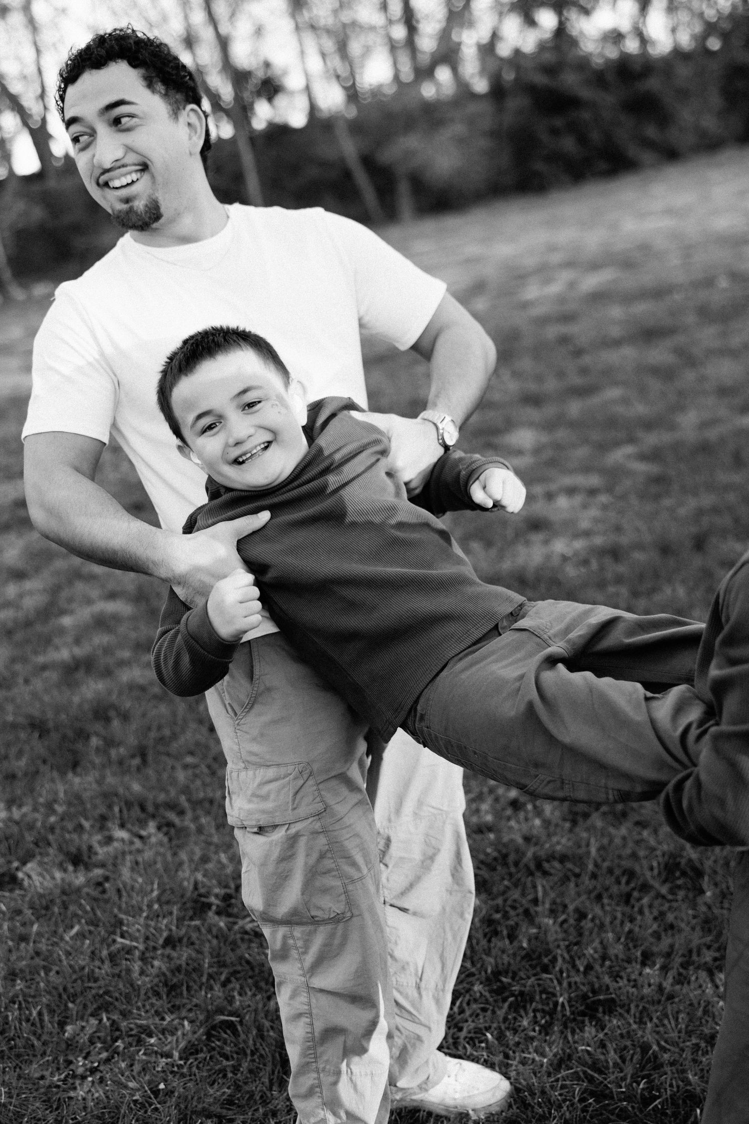 Father lifting and playing with his smiling son outdoors in a park in black and white photo.