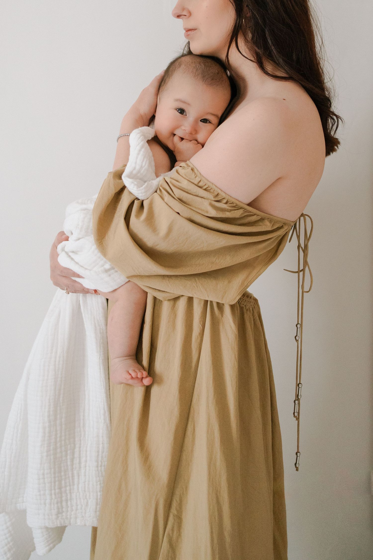 A tender moment captured as a baby in white dress nestles against someone wearing a flowing beige off-shoulder dress.