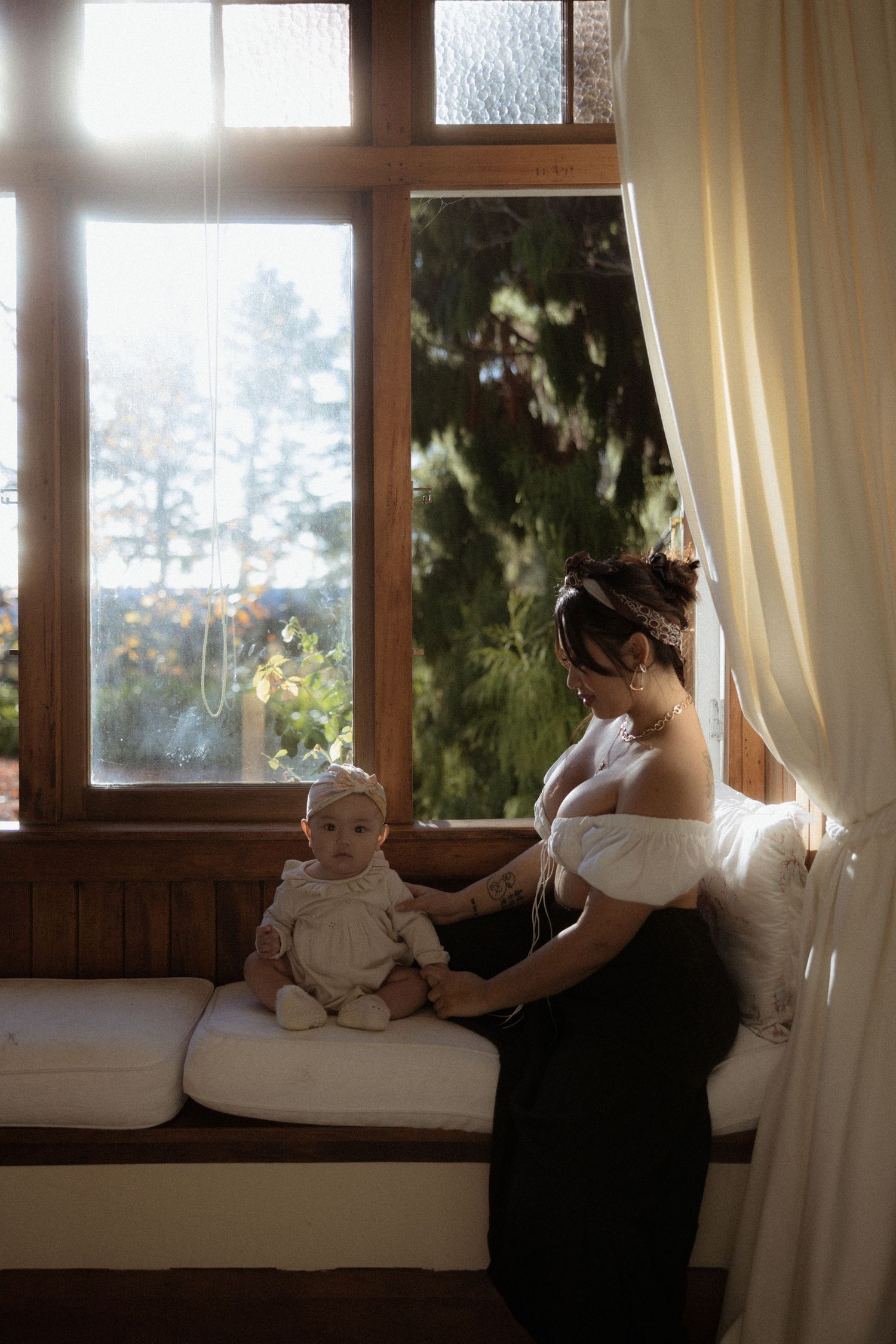 Woman in white off-shoulder top sits by window with baby doll on cushioned bench in moody lighting.