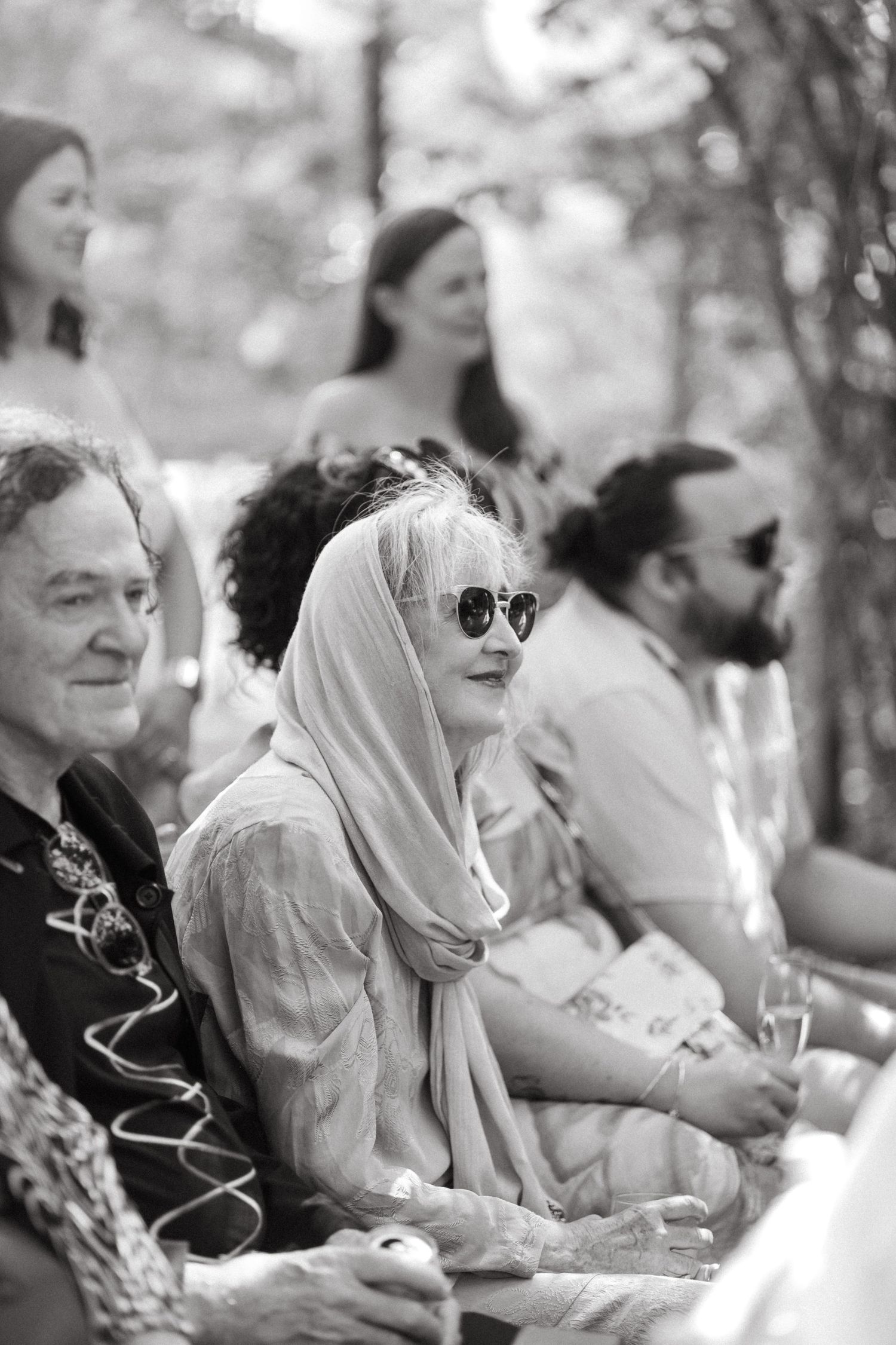 Black and white photo of wedding guests seated outdoors wearing sunglasses and head coverings.