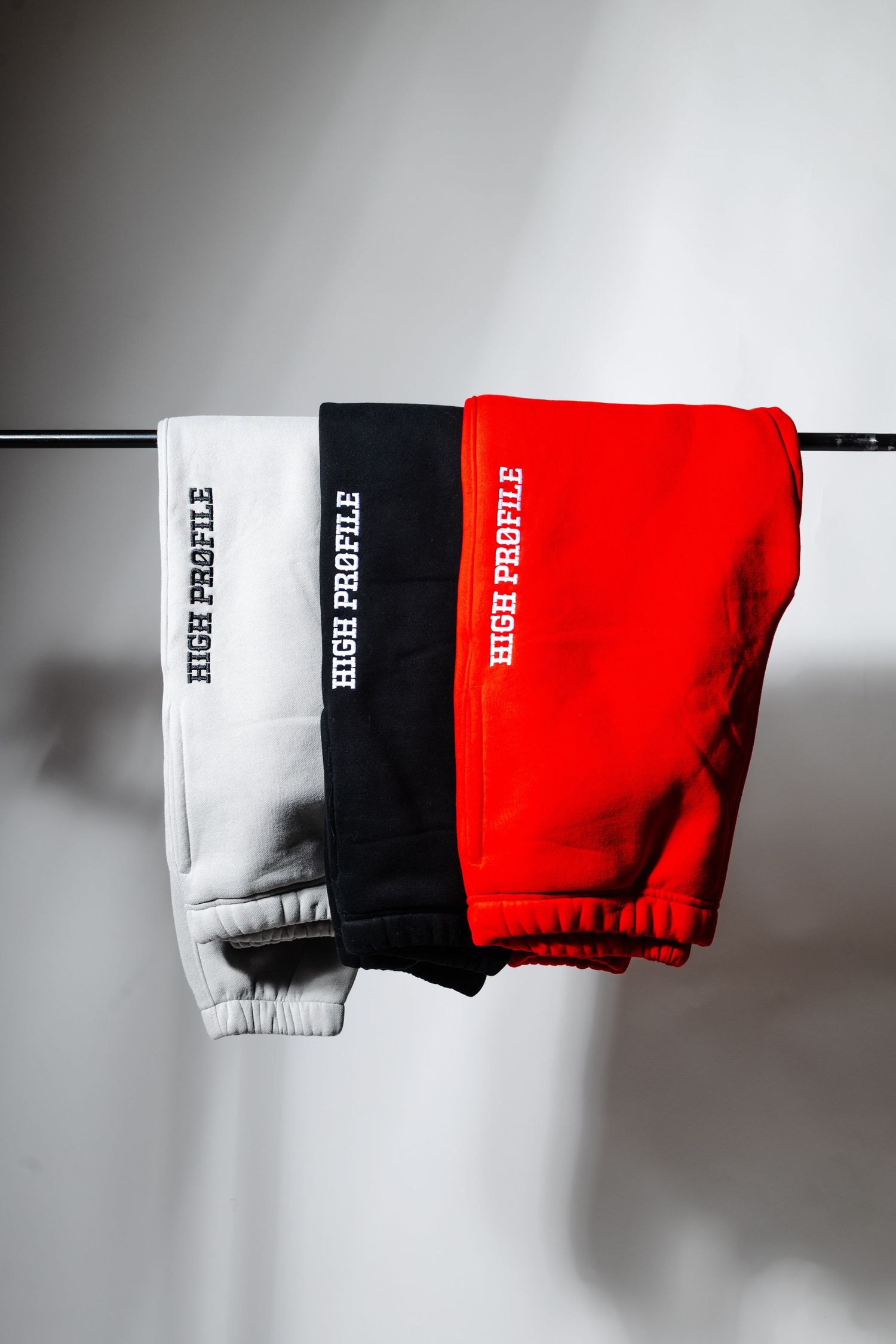 Three sweatpants in white, black and red hanging on clothing rack against light grey background.