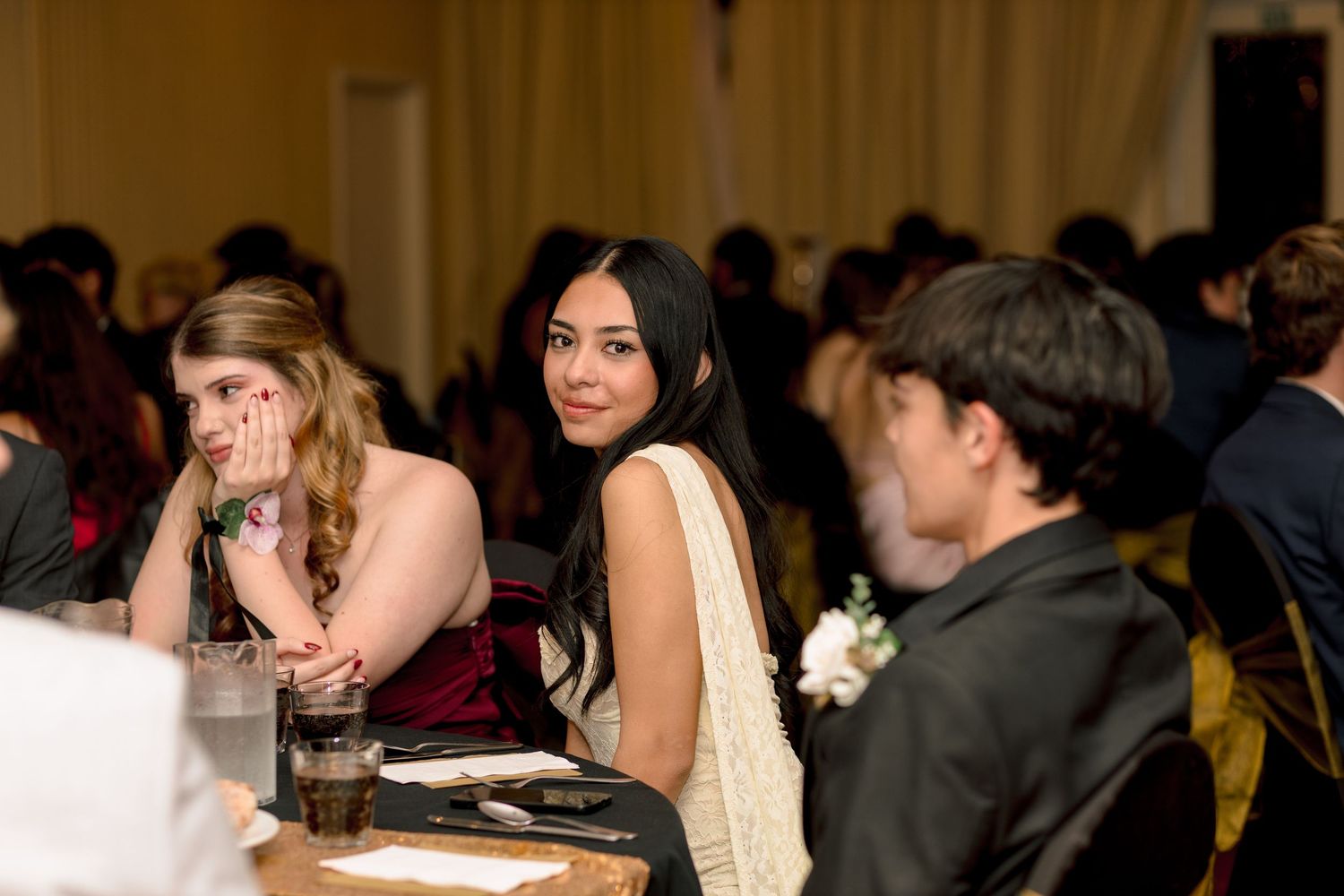 Teens dressed in formal attire socialize at a prom or banquet dinner event in an elegant venue.