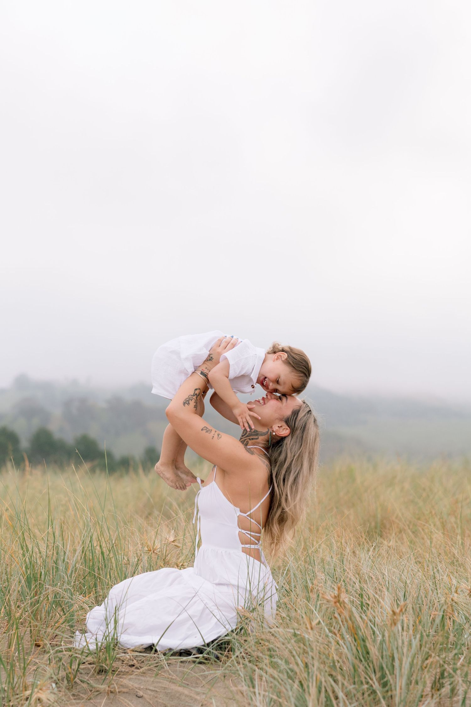 A mother in a white dress lifts and embraces her baby in a grassy field on a misty day.