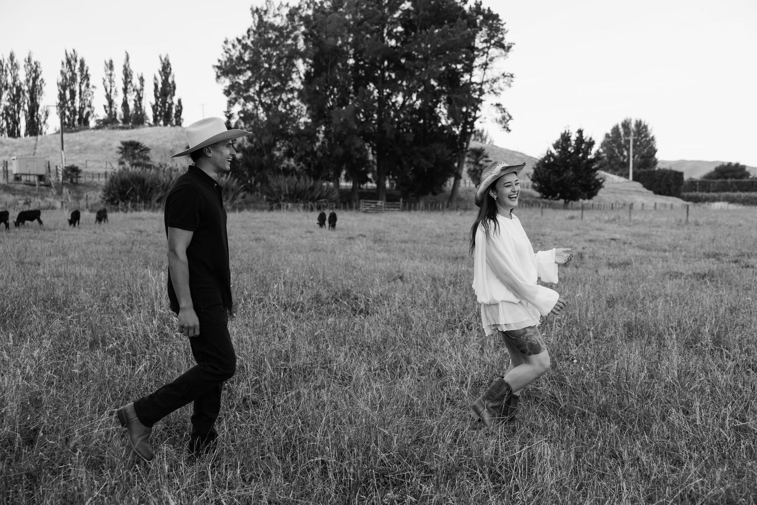 Black and white photo of a couple walking through a meadow with trees in the distance.