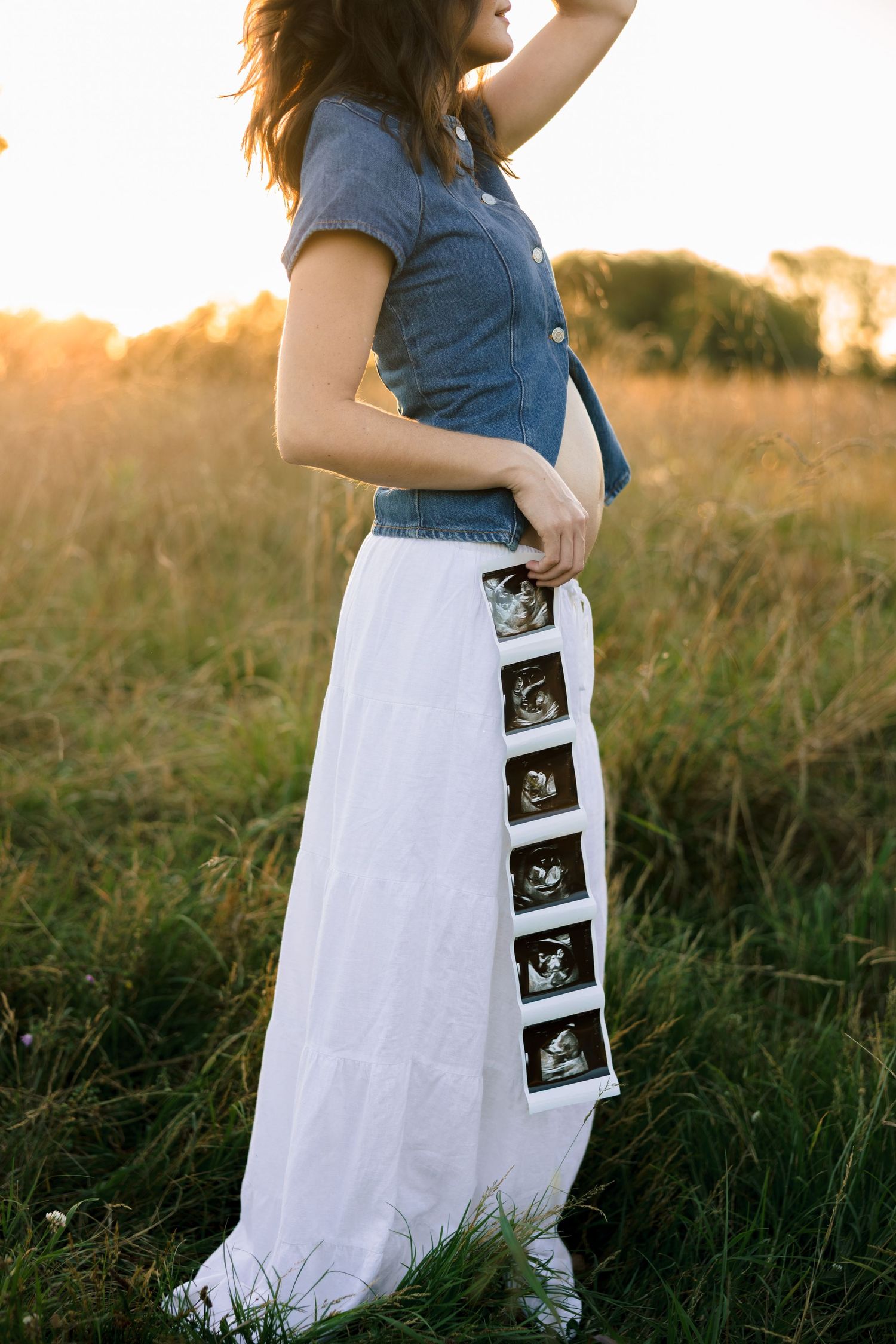 A pregnancy announcement photo series shows ultrasound images arranged on a flowing white dress against tall grass at sunset.