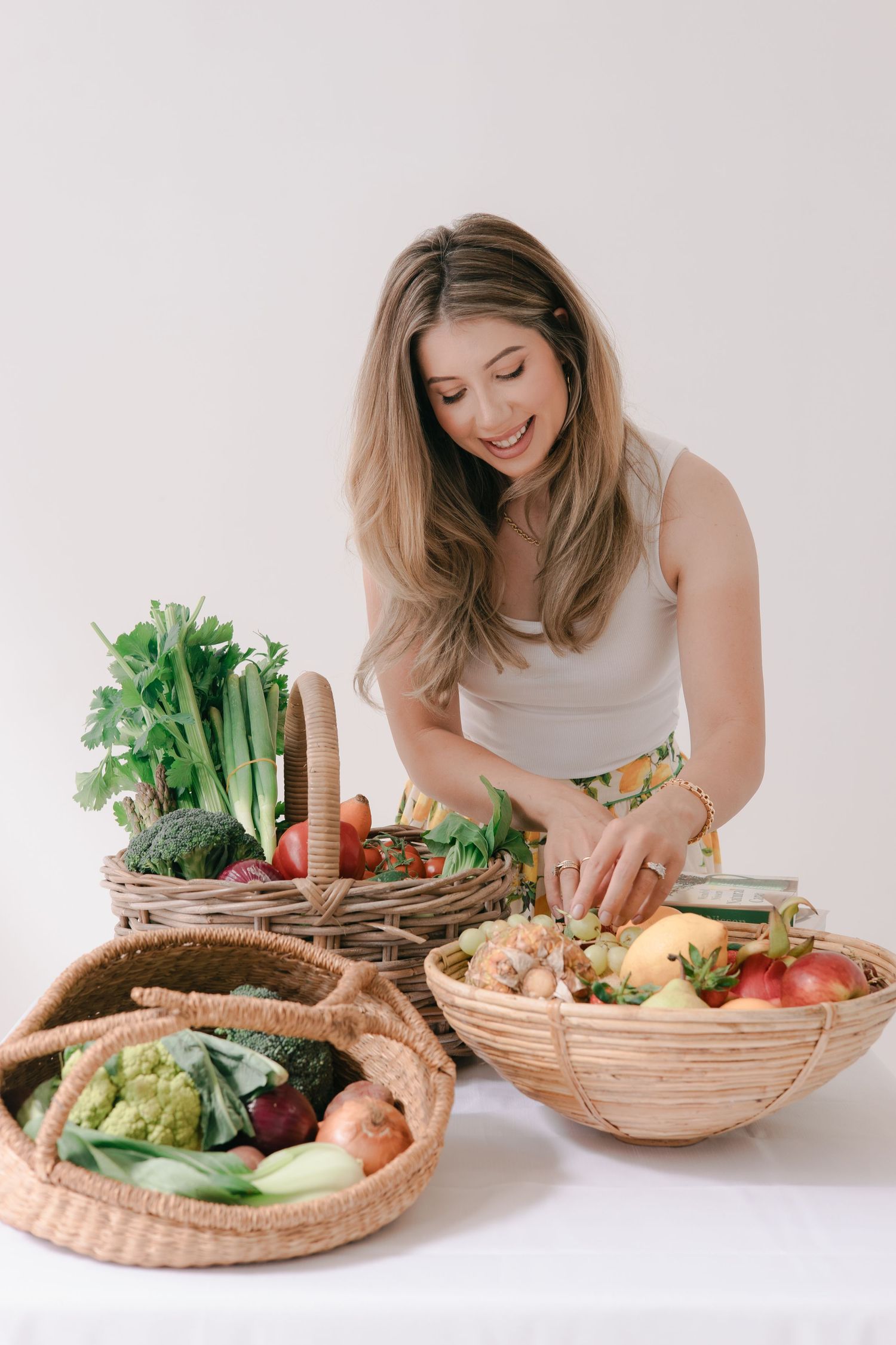 A woman in a white tank top arranges fresh vegetables and dips in woven baskets on a white surface.