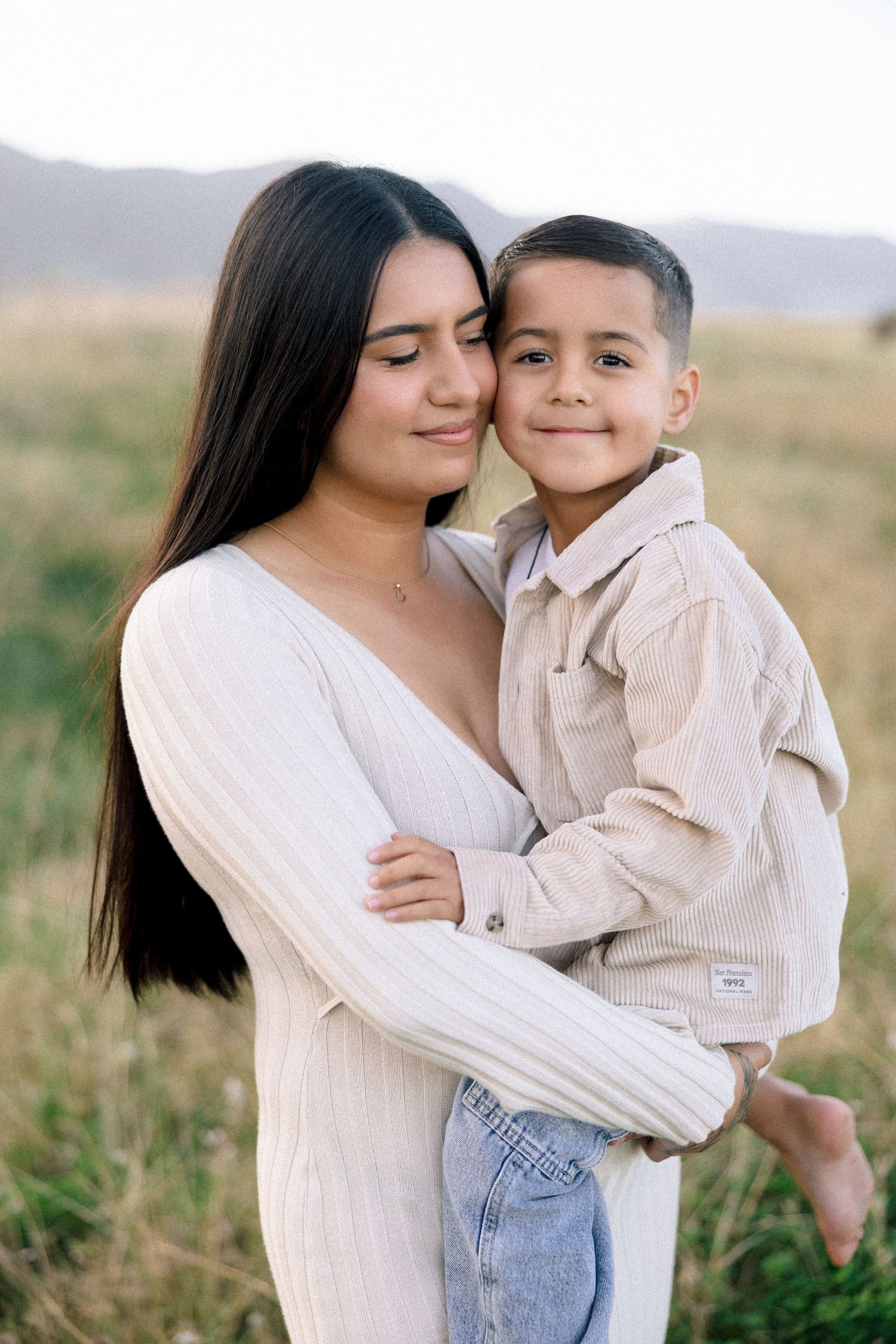 A young woman in a white sweater tenderly holds a smiling boy in a beige shirt outdoors.