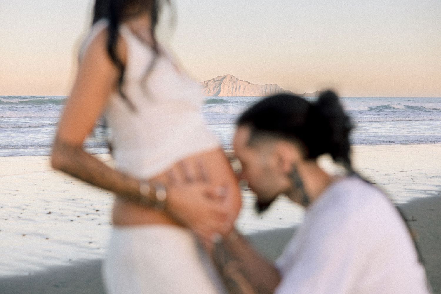 Couple at beach maternity shoot, man kissing pregnant woman's belly at sunset with ocean waves in background.