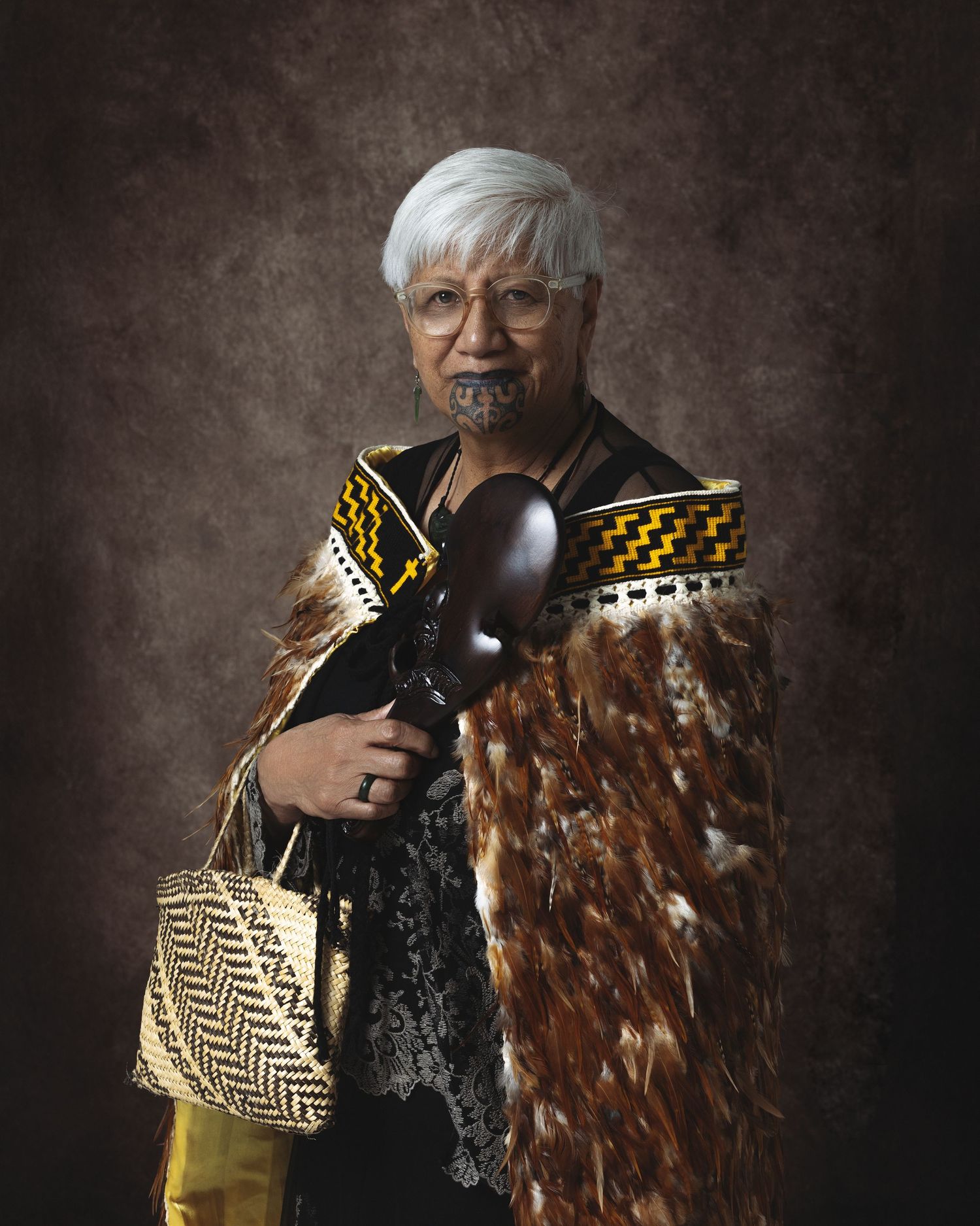 Maori elder in traditional korowai cloak with ta moko facial tattoo, holding a carved wooden weapon and woven kete bag.