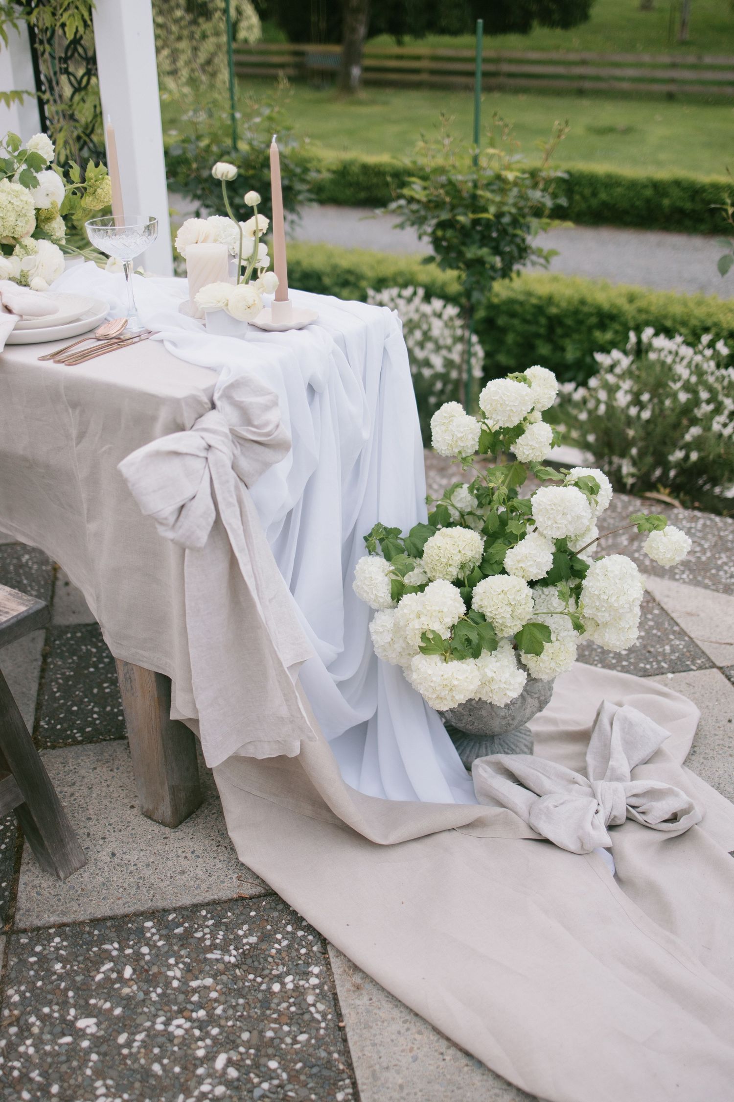 Elegant wedding table setting with white hydrangeas and linen bows on stone patio overlooking garden landscape.