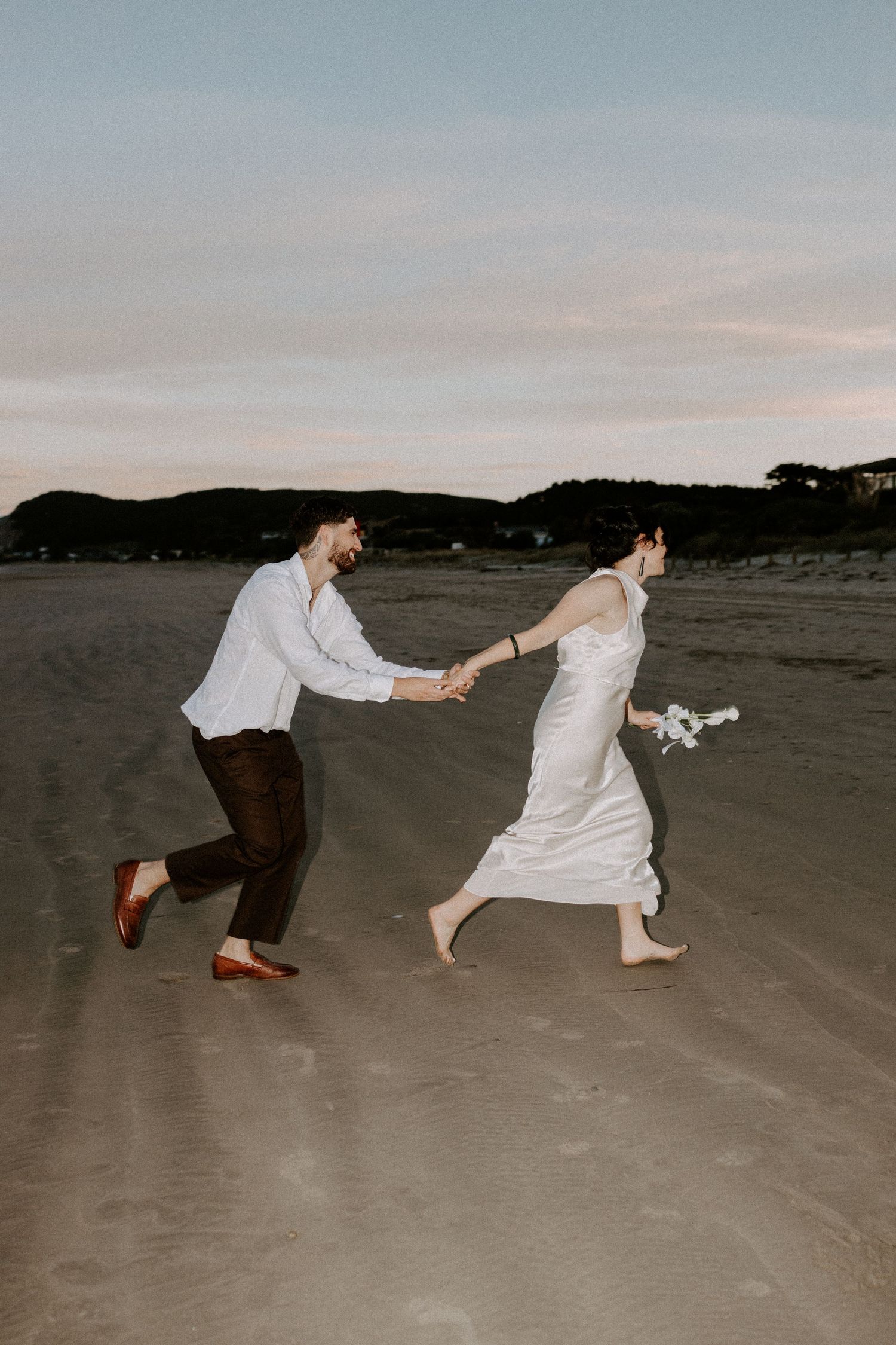 A playful couple runs and holds hands on an empty beach at dusk wearing white wedding attire.