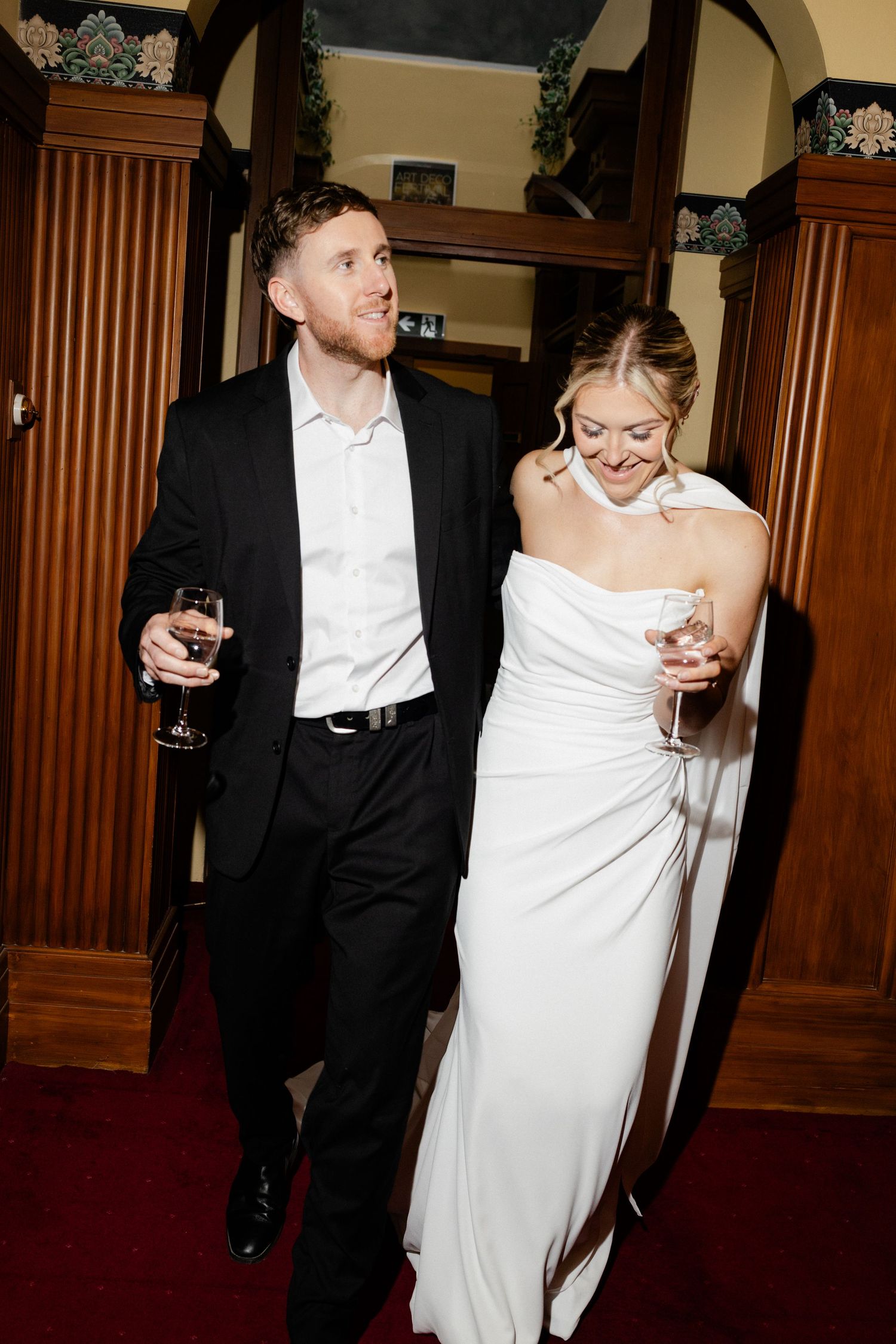 A couple in formal wedding attire share a laugh while holding wine glasses at an indoor reception.
