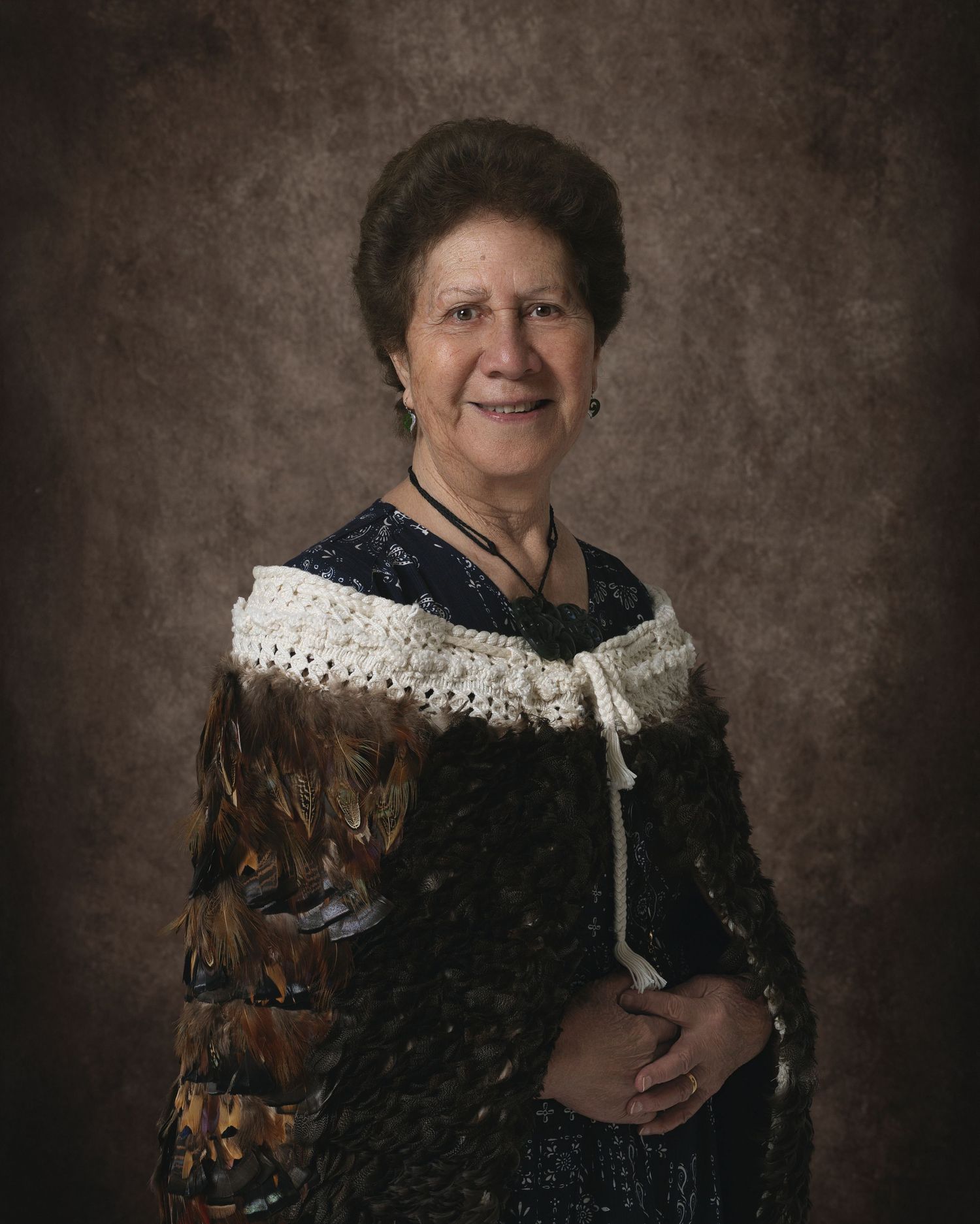 Elderly woman smiling in traditional Maori feather cloak and crochet collar, posed for a formal portrait.
