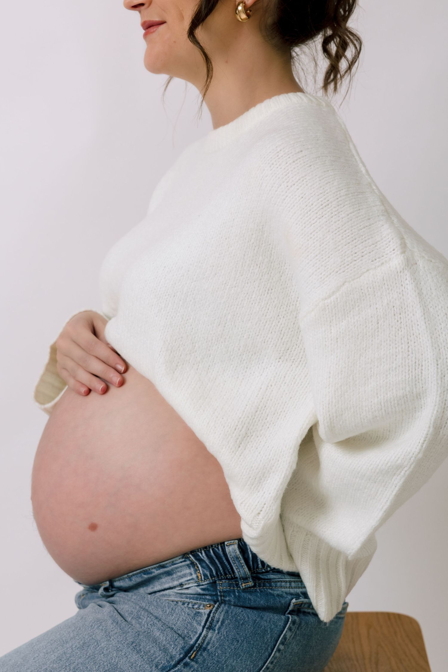 Pregnant woman in white knit sweater and jeans cradling her bare baby bump against a white background.