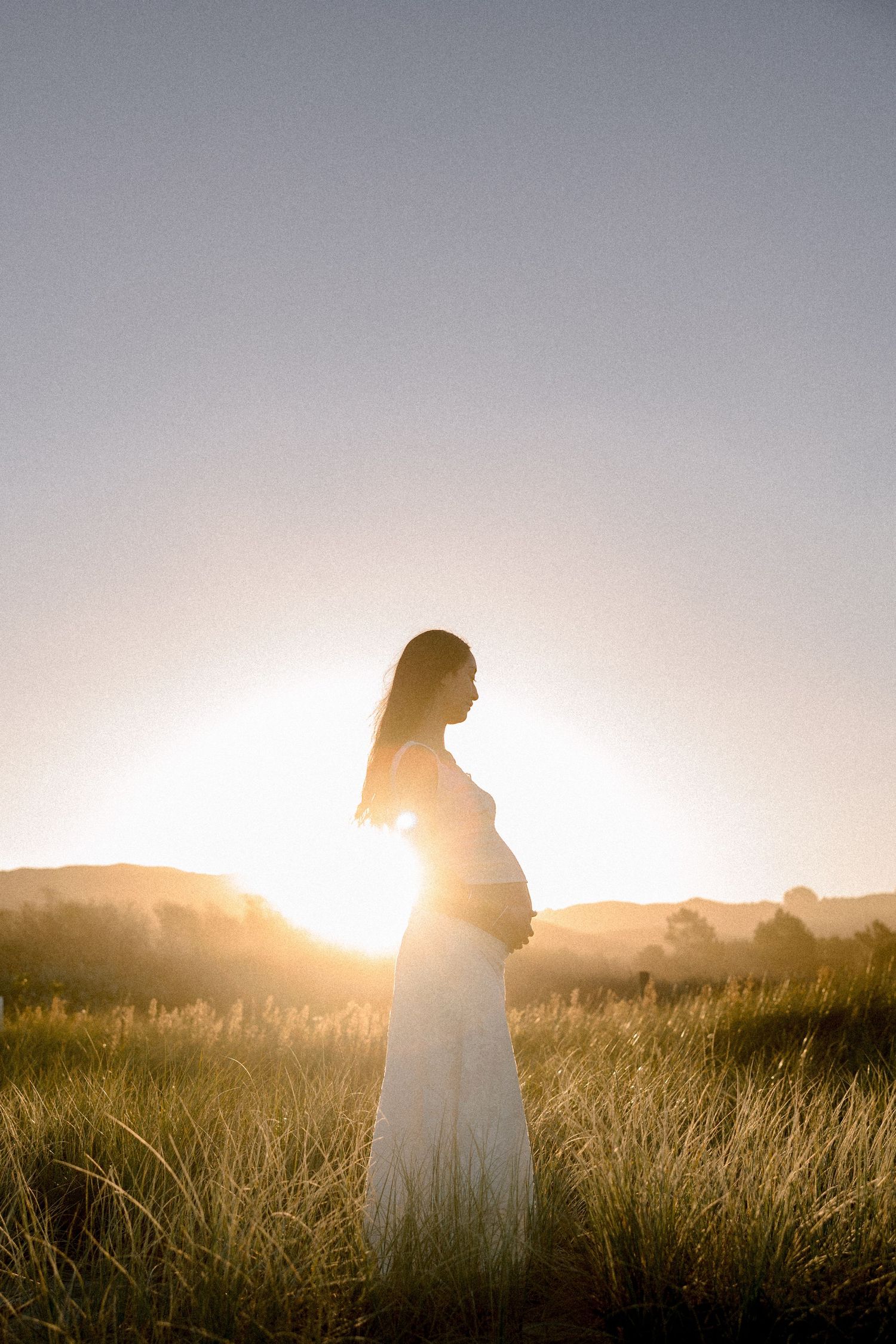 Pregnant woman in white dress standing in golden field at sunset, silhouetted against warm glowing light.