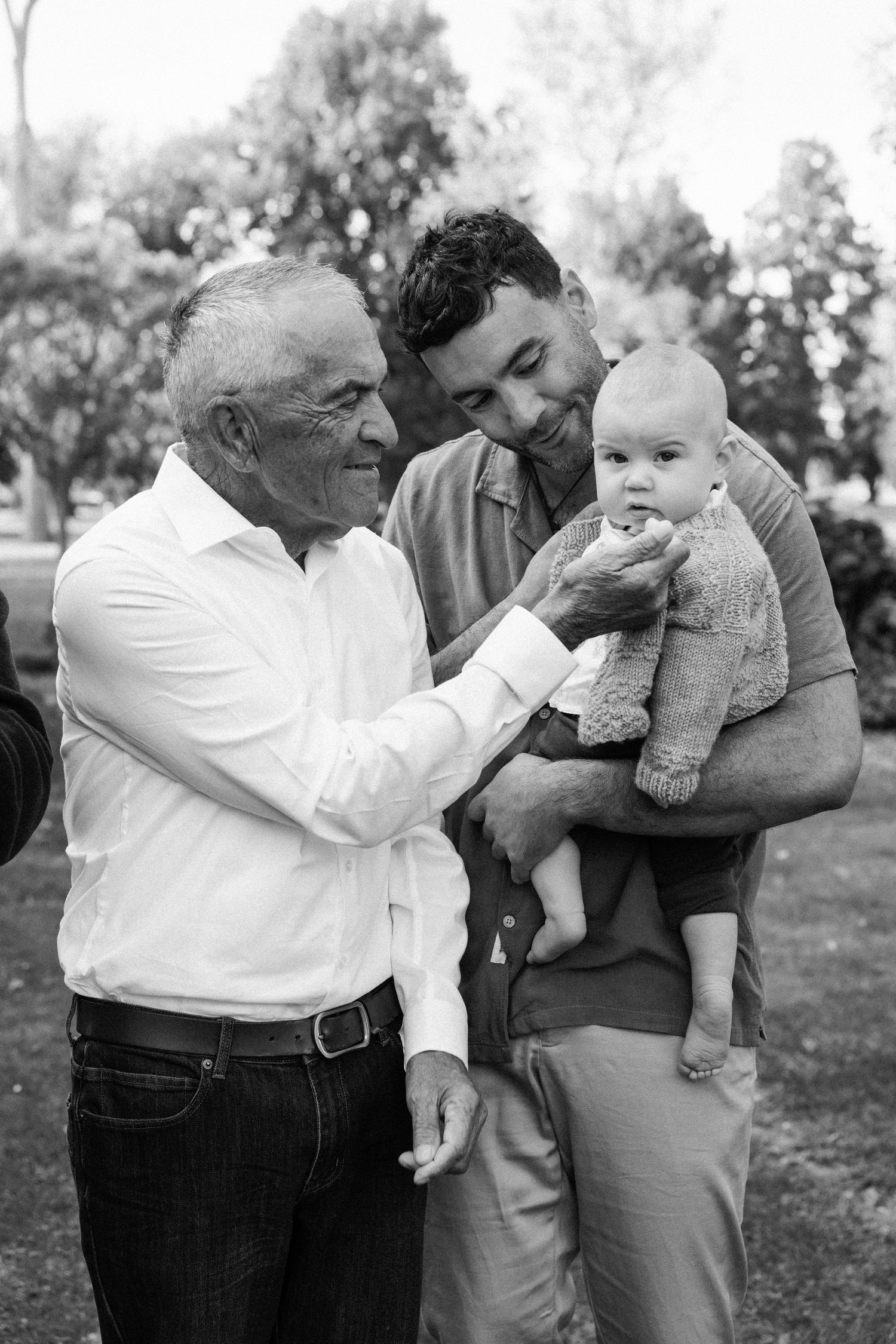 Black and white photo of three generations, grandfather touching baby held by father in a park.