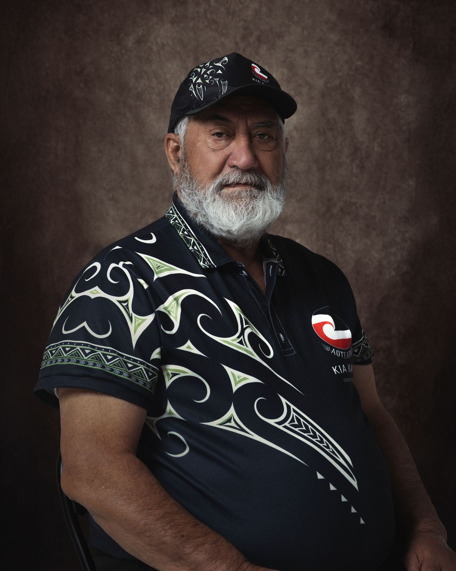 Elderly Maori man with white beard wearing traditional Maori patterned shirt and cap, posed for a formal portrait.