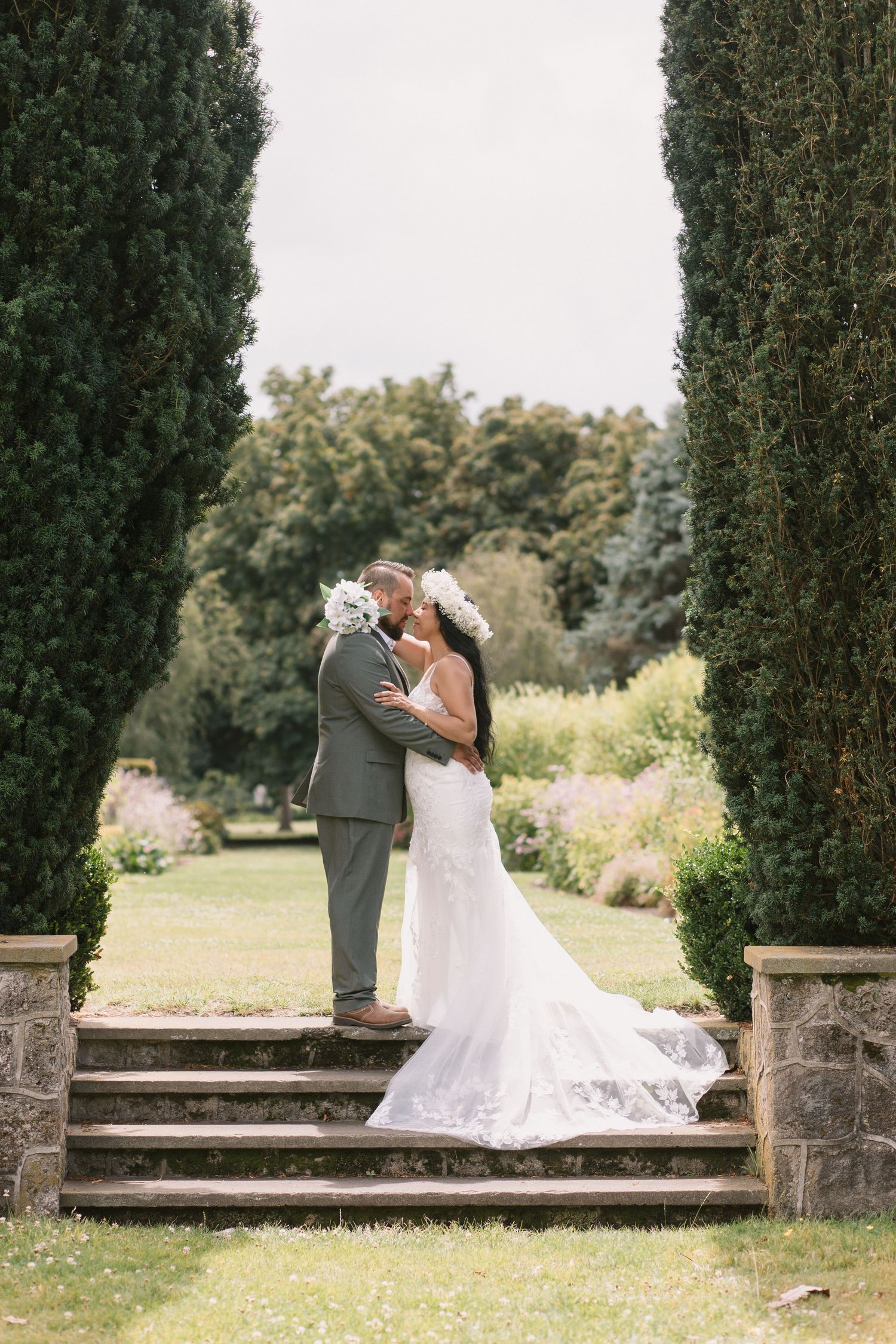 A wedding couple shares a romantic kiss on stone steps between tall cypress trees in a garden setting.