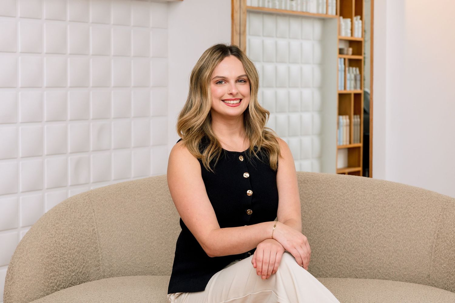 Smiling young woman with blonde hair sitting on a beige sofa in a modern white interior space.