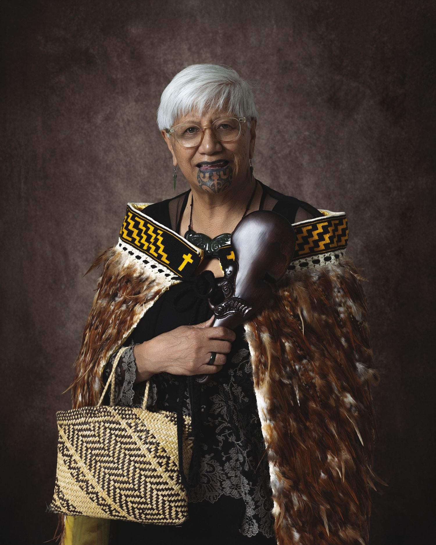 Maori elder woman in traditional feather cloak, ta moko facial tattoo, holding carved weapon and woven flax kete basket.