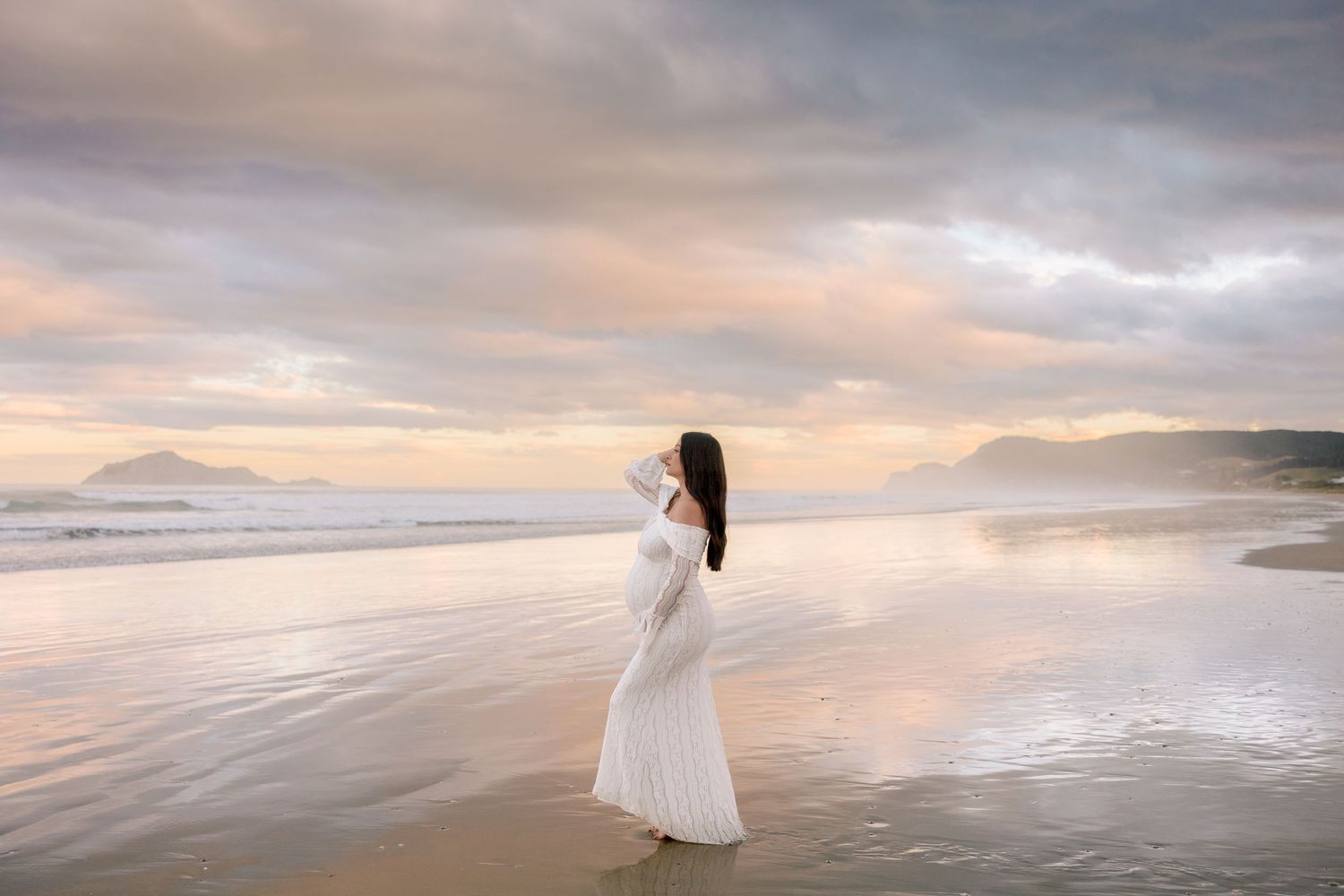 A silhouette stands in a flowing white dress on a misty beach at sunset with mountains in the background.